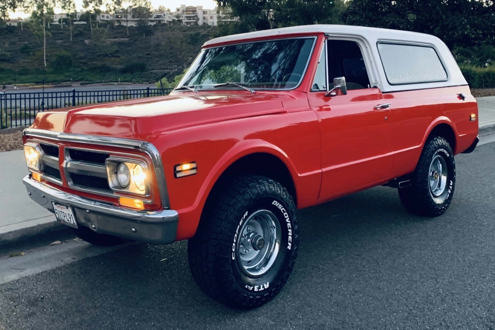 Gmc Jimmy 桌布 壁紙 — 一輛鮮紅色經典雪佛蘭K5 Blazer，白色車頂，停泊在黃昏時分靠近圍欄和綠色山丘的瀝青路上。