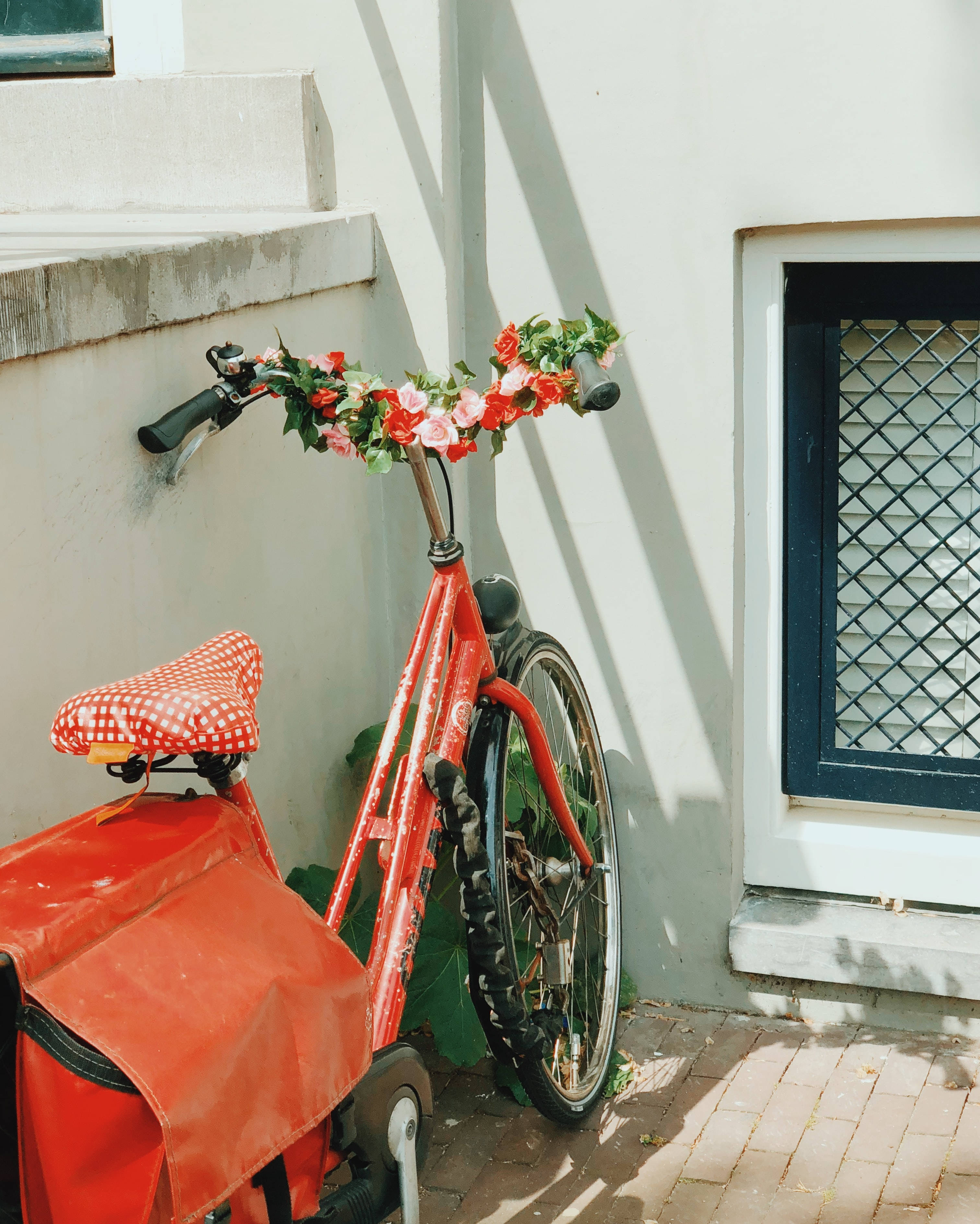 Bicycle 桌布 壁紙 — 一輛鮮紅色自行車飾有花環，靠在陽光照射的白牆旁，旁邊是一條磚砌小徑。
