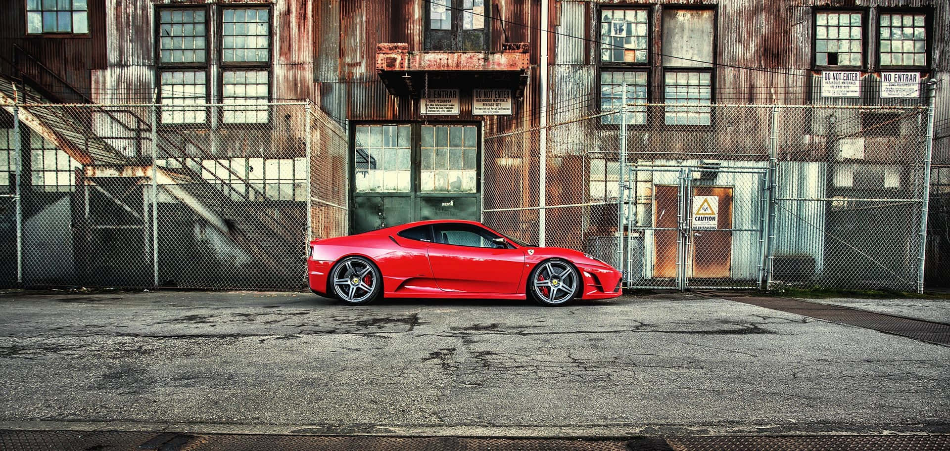 Ferrari F430 桌布 壁紙 — 一輛鮮紅色跑車停在一棟風化的工業建築前，建築物有生鏽的金屬和鐵絲網圍欄。