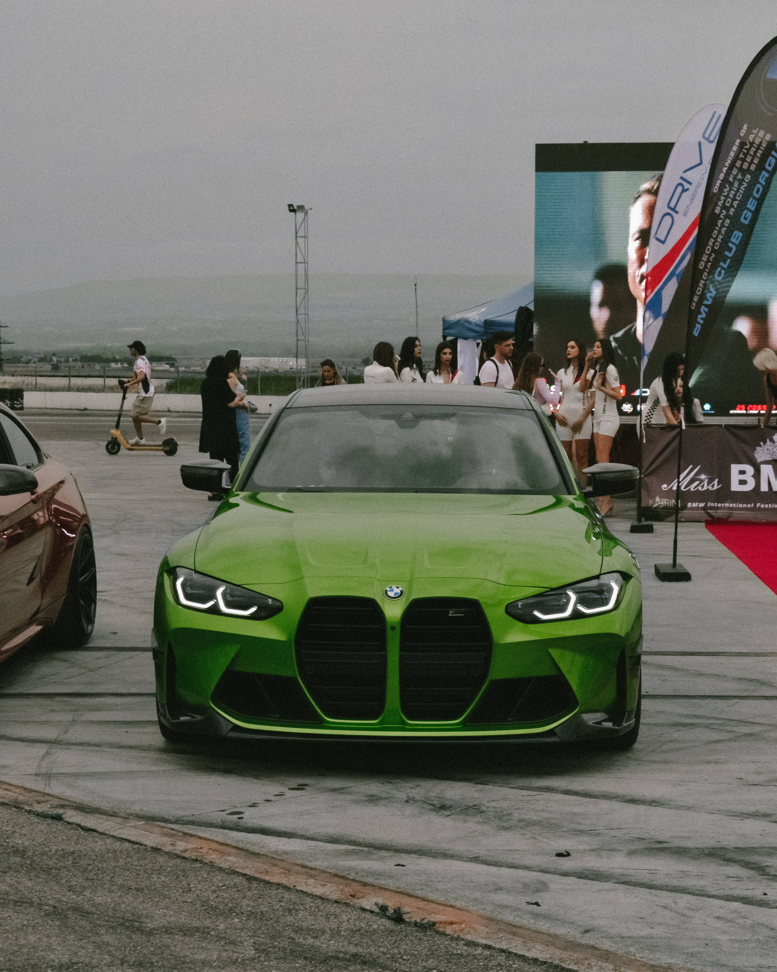 Racing 桌布 壁紙 — 一輛鮮綠色BMW M3跑車停放在潮濕的瀝青停車場，背景有人群和活動橫幅，天空多雲。
