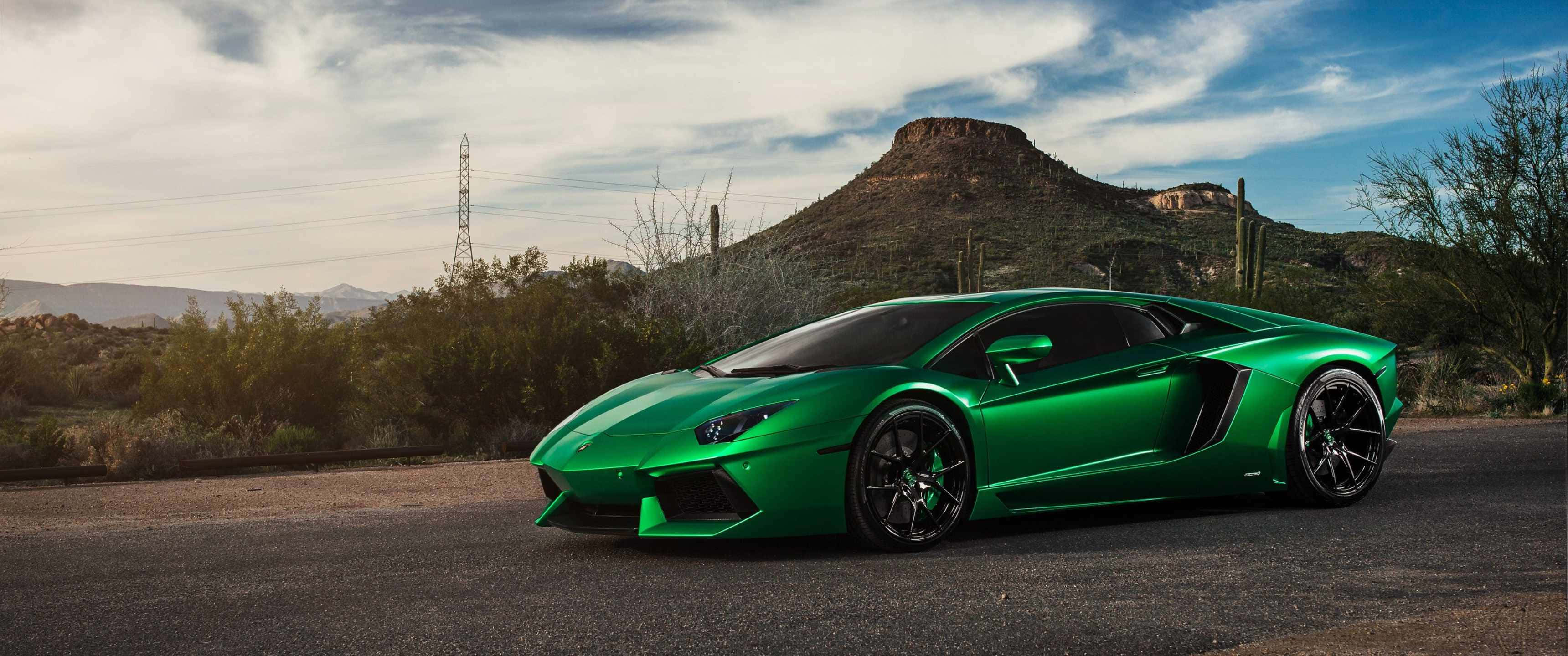 3440x1440p Lamborghini 桌布 壁紙 — 一輛鮮綠色藍寶堅尼Aventador停泊在沙漠道路上，周圍有仙人掌和岩石山丘，天空部分多雲。