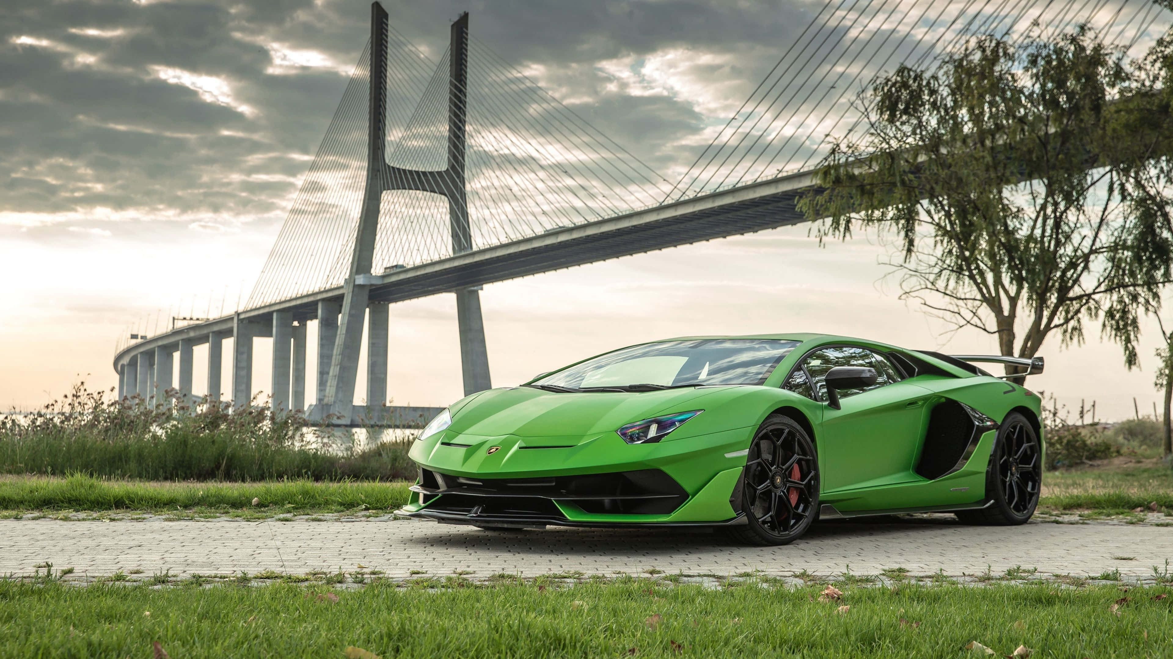 4K 藍寶堅尼 桌布 壁紙 — 一輛鮮綠色藍寶堅尼Aventador SVJ停放在鋪砌小徑上，背後一座巨大的斜張橋在陰天下聳立。