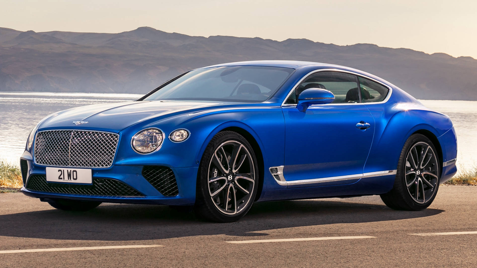 bentley HD 桌布 壁紙 — 一輛鮮藍色 Bentley Continental GT 轎跑車在金色陽光下閃閃發光，停泊在平靜的海濱，遠處有山丘。