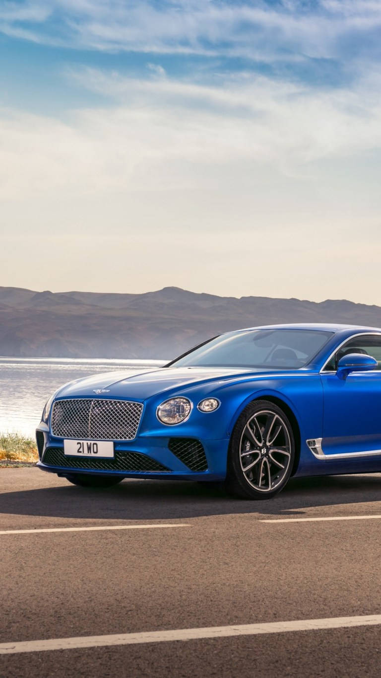 手機 桌布 壁紙 — 一輛鮮藍色 Bentley Continental GT 雙門跑車停泊在沿海道路上，背景是山脈和海洋，天空部分多雲。