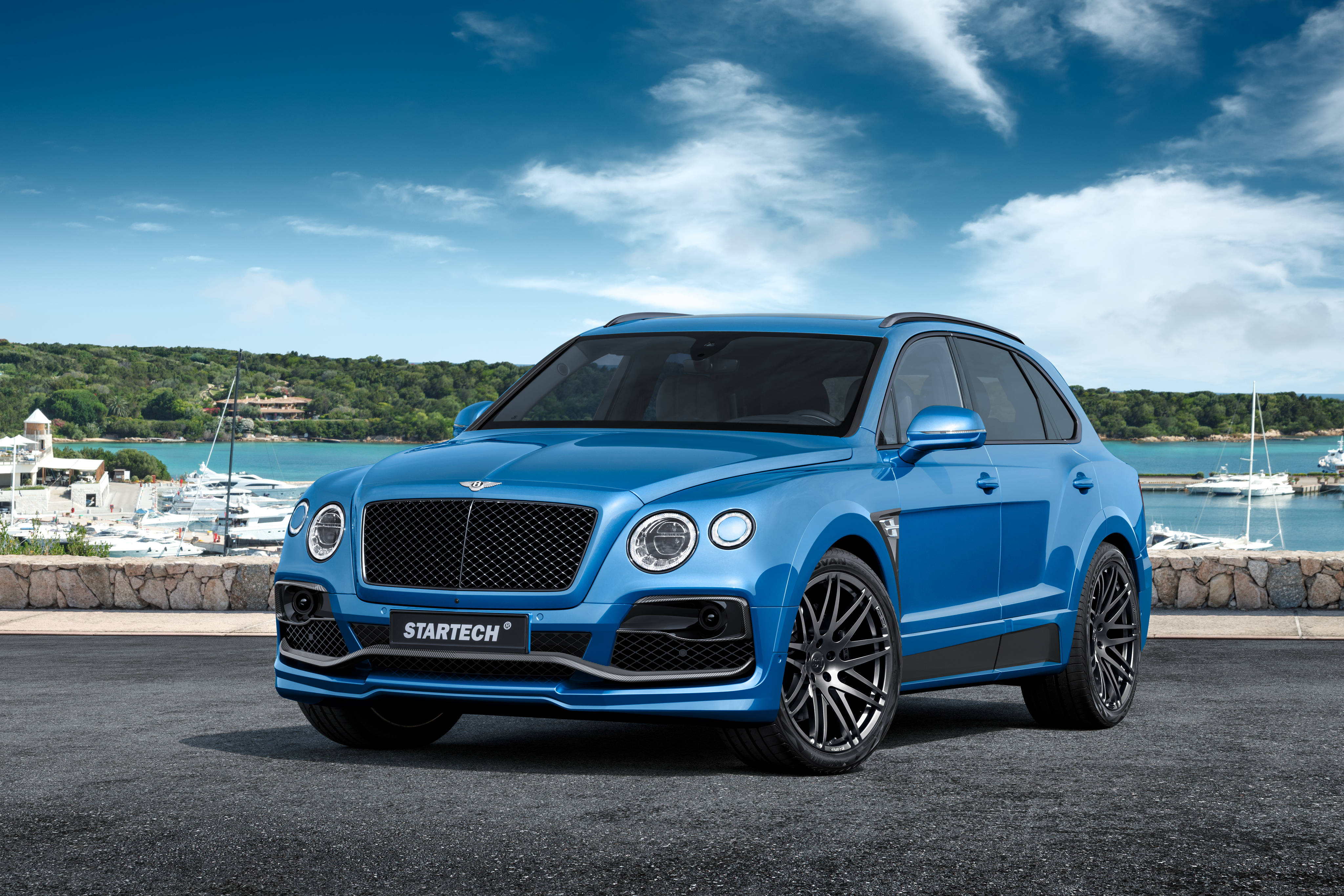 賓利 桌布 壁紙 — 一輛鮮藍色、經Startech改裝的Bentley Bentayga SUV停泊在沿海道路上，背景有遊艇和綠色山丘，天空湛藍。