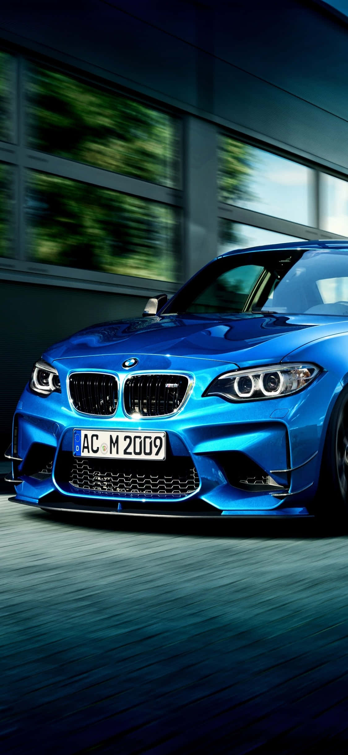 iPhone X Bmw 桌布 壁紙 — 一輛鮮藍色BMW M2跑車以動態模糊效果高速駛過一棟現代建築，強調其動感。