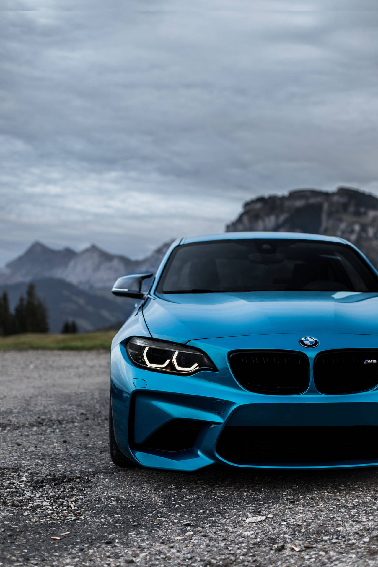 多雲天空下，藍色BMW M2跑車停泊在碎石上靠近山脈 — BMW 動圖 壁紙