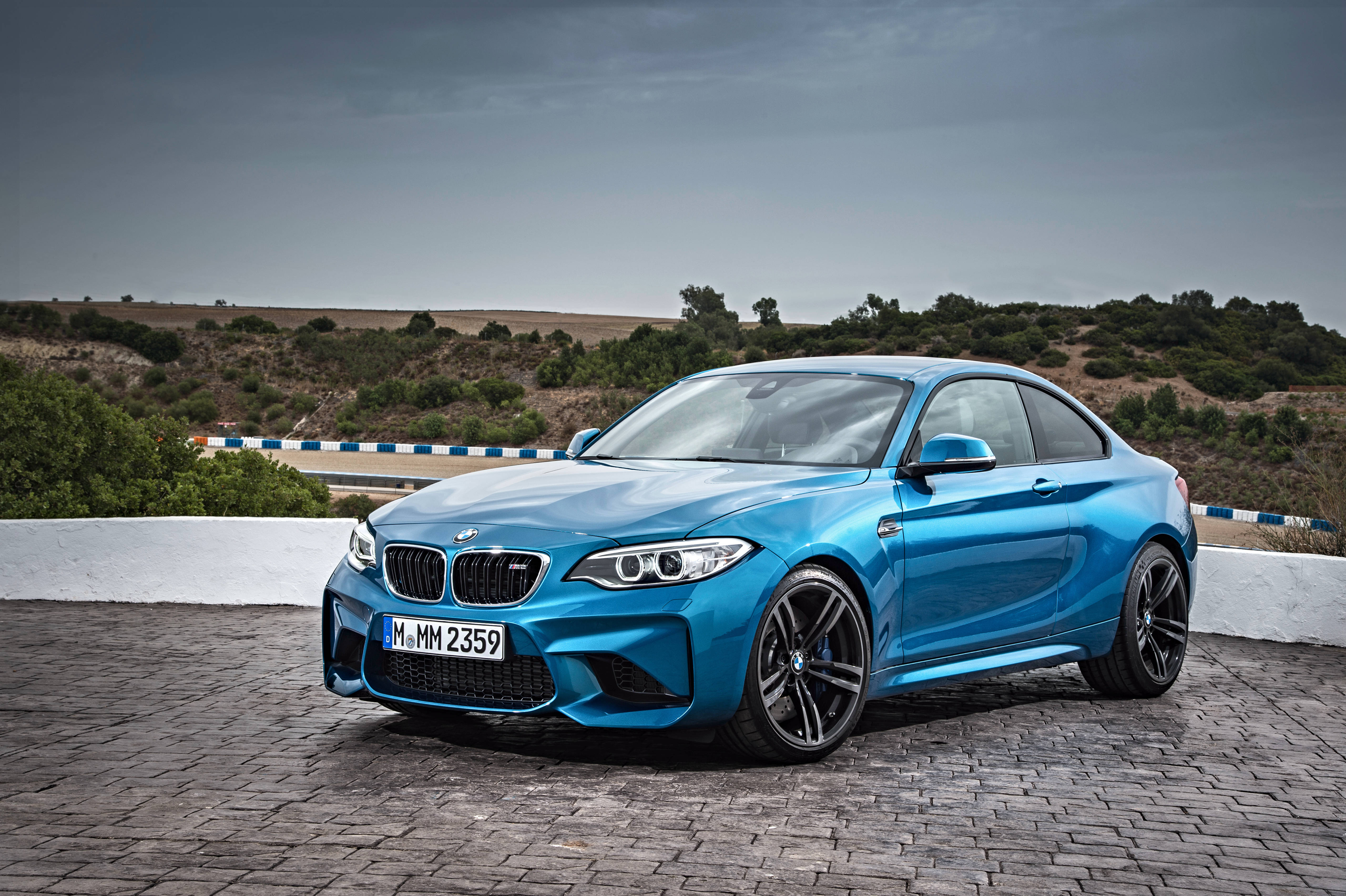 BMW 動圖 桌布 壁紙 — 一輛鮮藍色BMW M2雙門跑車停在鵝卵石觀景點，背景有起伏的山丘和陰天。