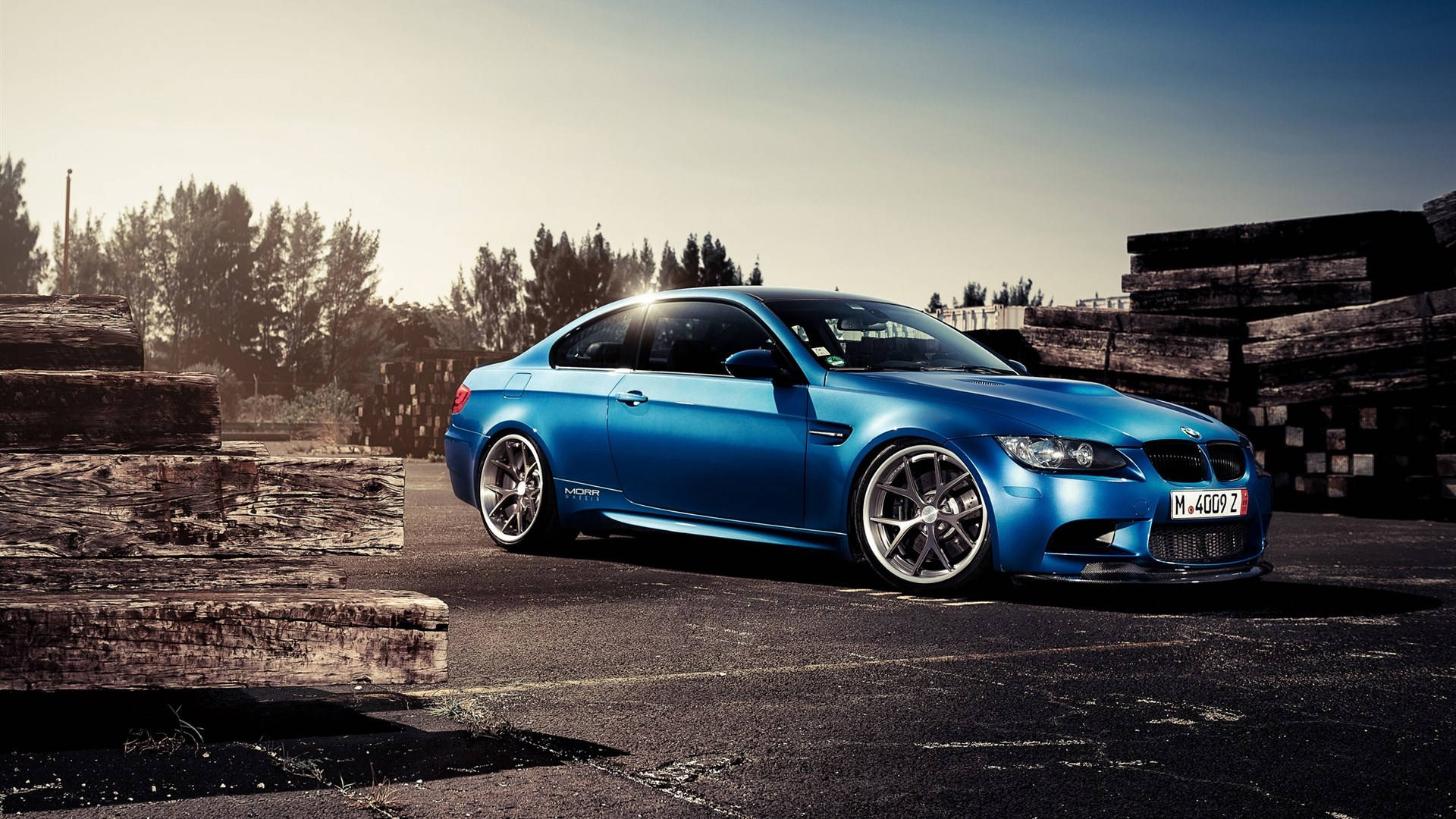 桌布 壁紙 — 一輛鮮藍色BMW M3雙門跑車停在瀝青路上，旁邊是堆疊的風化木頭，天空晴朗。