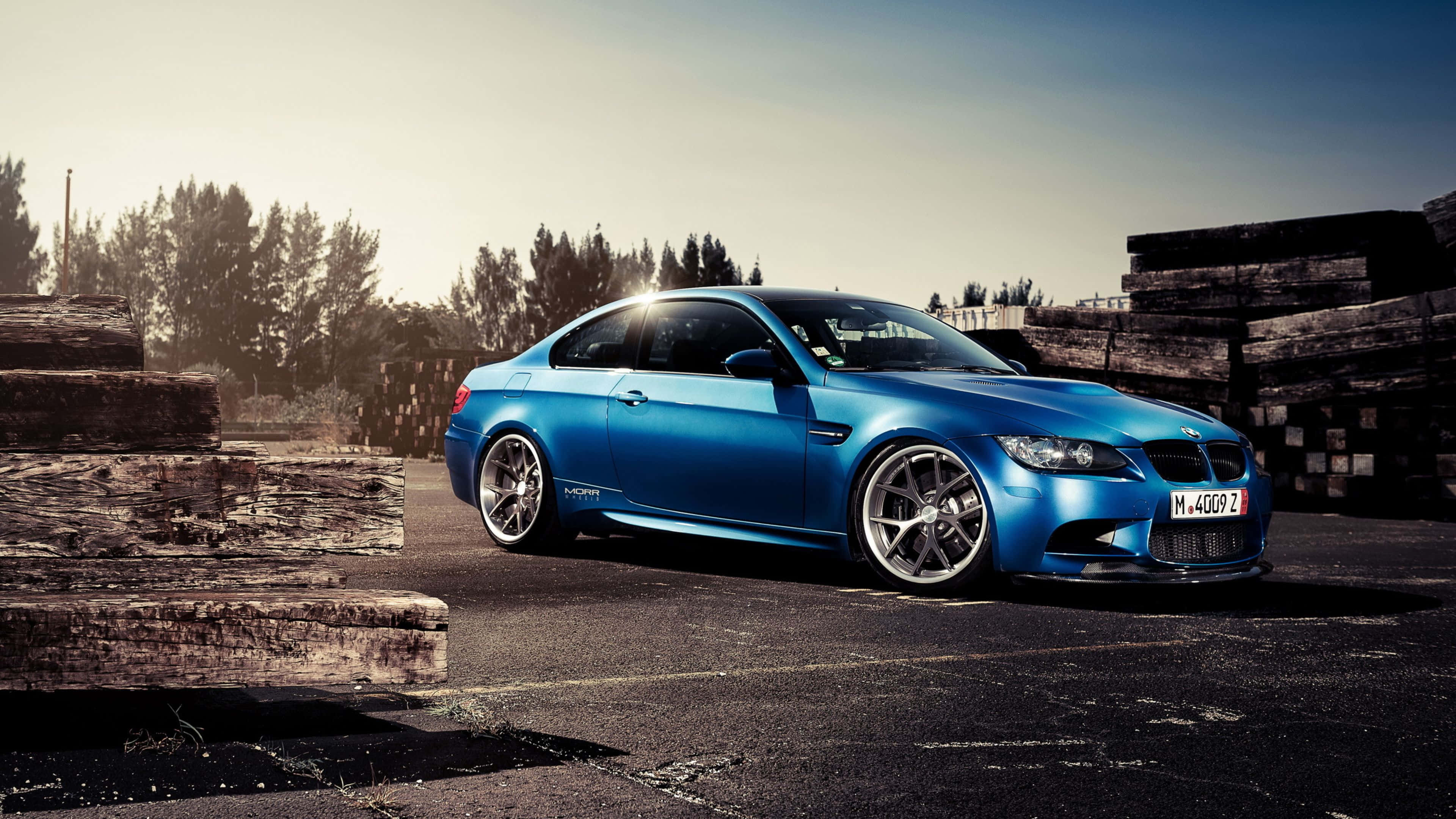 4K BMW 壁紙 桌布 壁紙 — 一輛鮮藍色BMW M3雙門跑車停在柏油路上，旁邊是堆疊的木頭板子，天空晴朗。