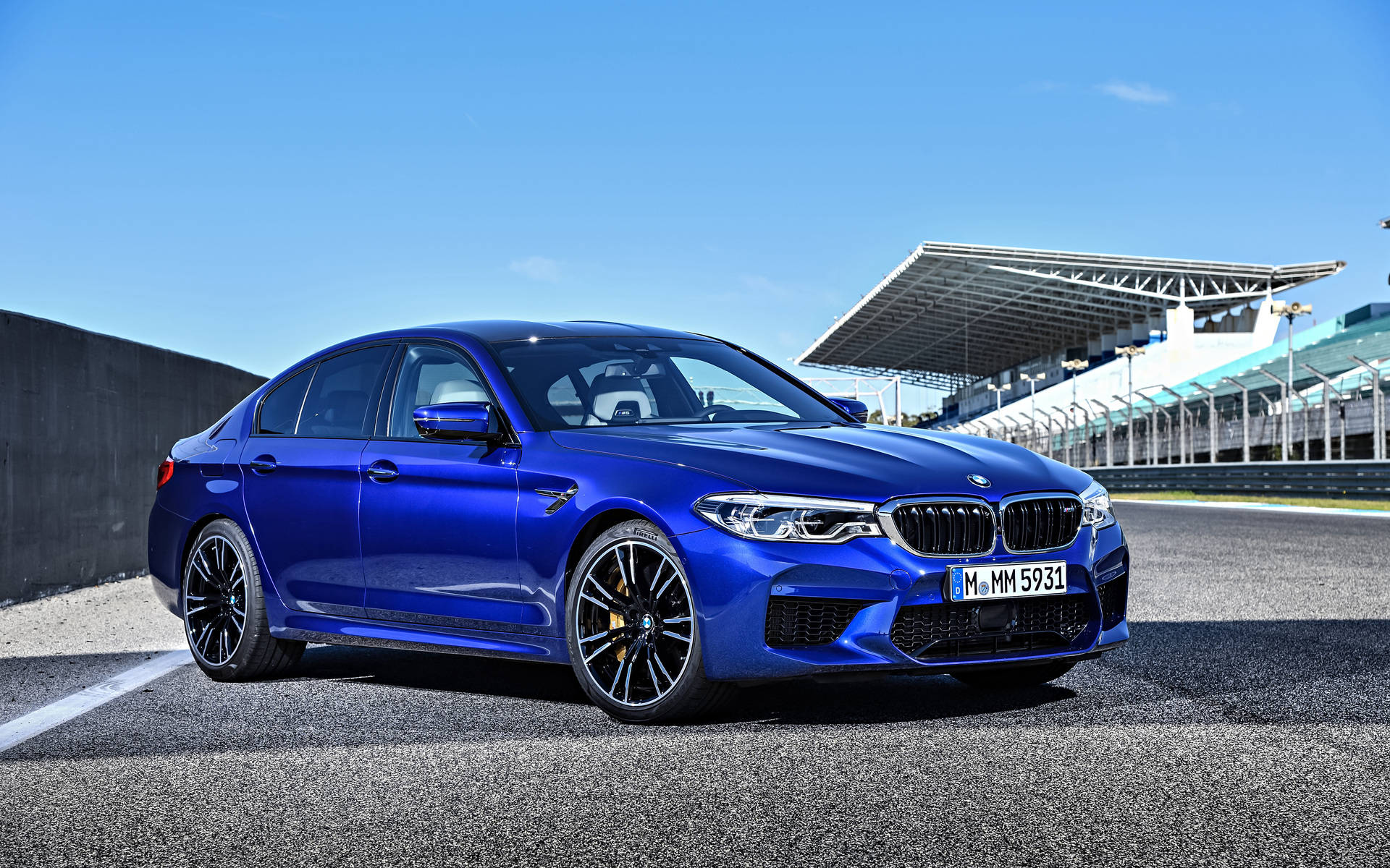 BMW 動圖 桌布 壁紙 — 一輛鮮藍色BMW M5轎車在明亮的陽光下，在瀝青賽道上閃閃發光，背景有看台。
