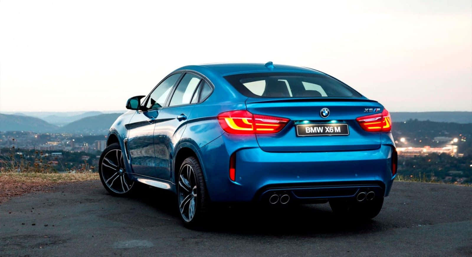 Bmw X6 M 桌布 壁紙 — 一輛鮮藍色BMW X6 M轎跑車停在俯瞰城市景觀的山頂上，在黃昏時分亮起尾燈。