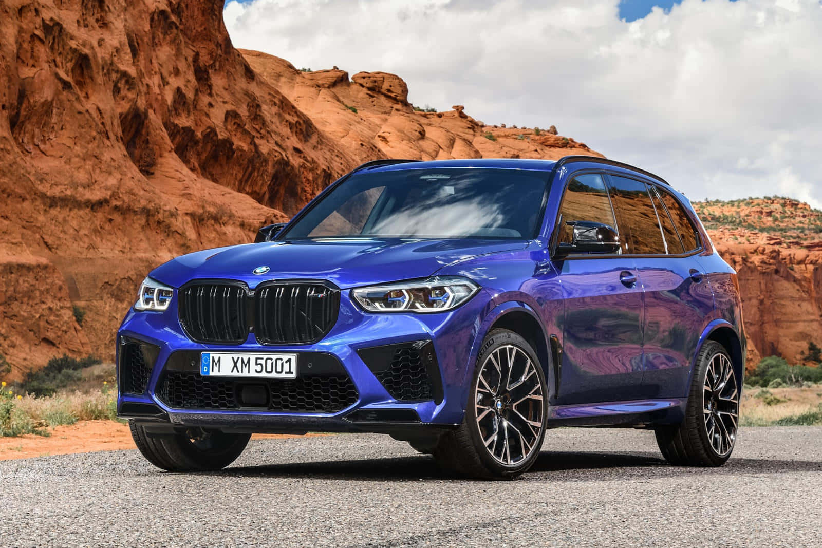 Bmw X5 桌布 壁紙 — 一輛鮮藍色BMW XM SUV停泊在碎石上，背景有紅色岩層和陰天。