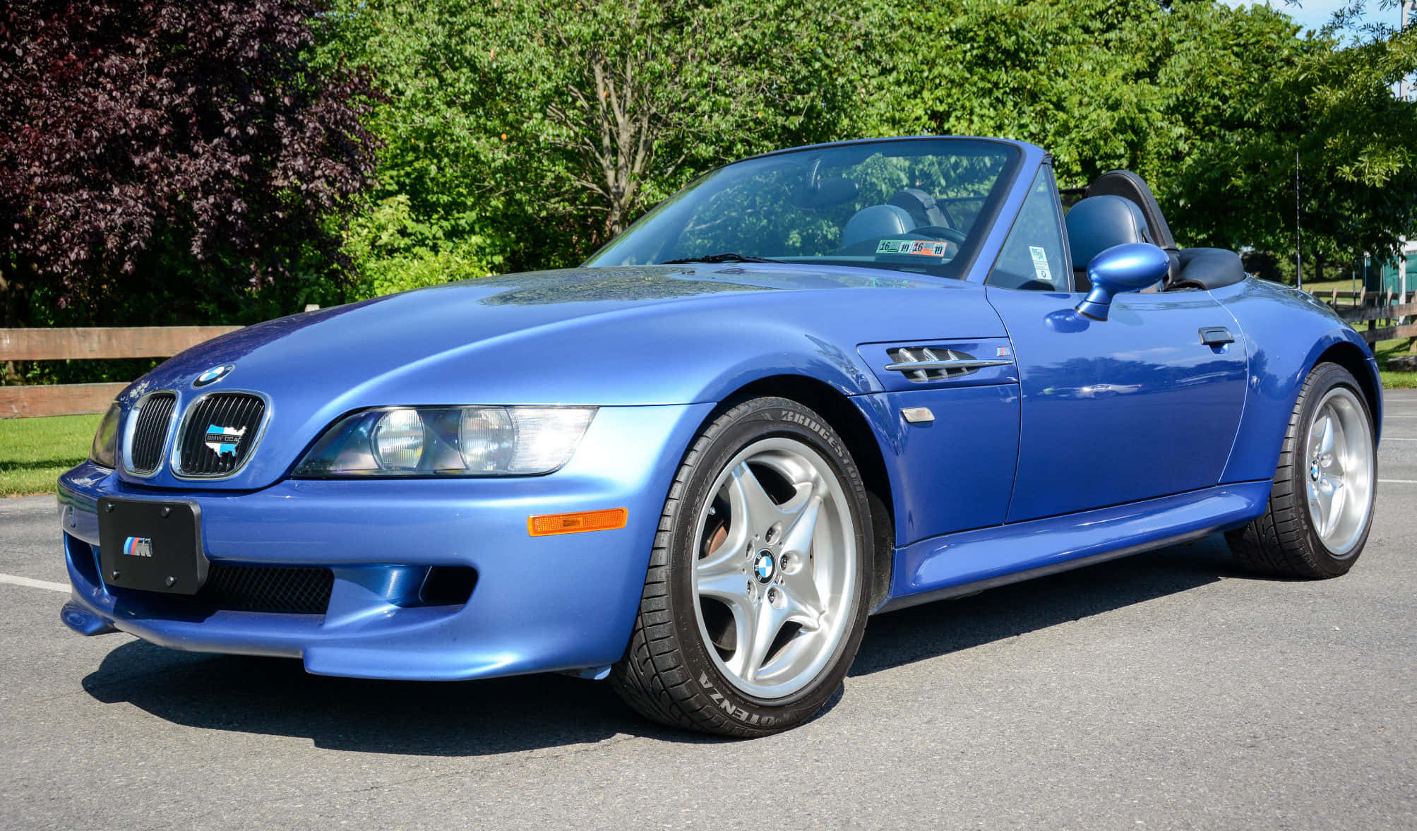 Bmw Z3 桌布 壁紙 — 一輛鮮藍色BMW Z8敞篷車停在柏油路上，背景有茂密的綠樹，在明亮的日光下。