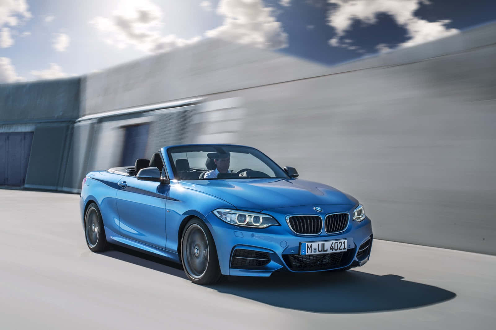 BMW 動圖 桌布 壁紙 — 一輛鮮藍色BMW敞篷車在晴朗天空下，沿著現代混凝土道路飛馳，天空有零星雲朵。