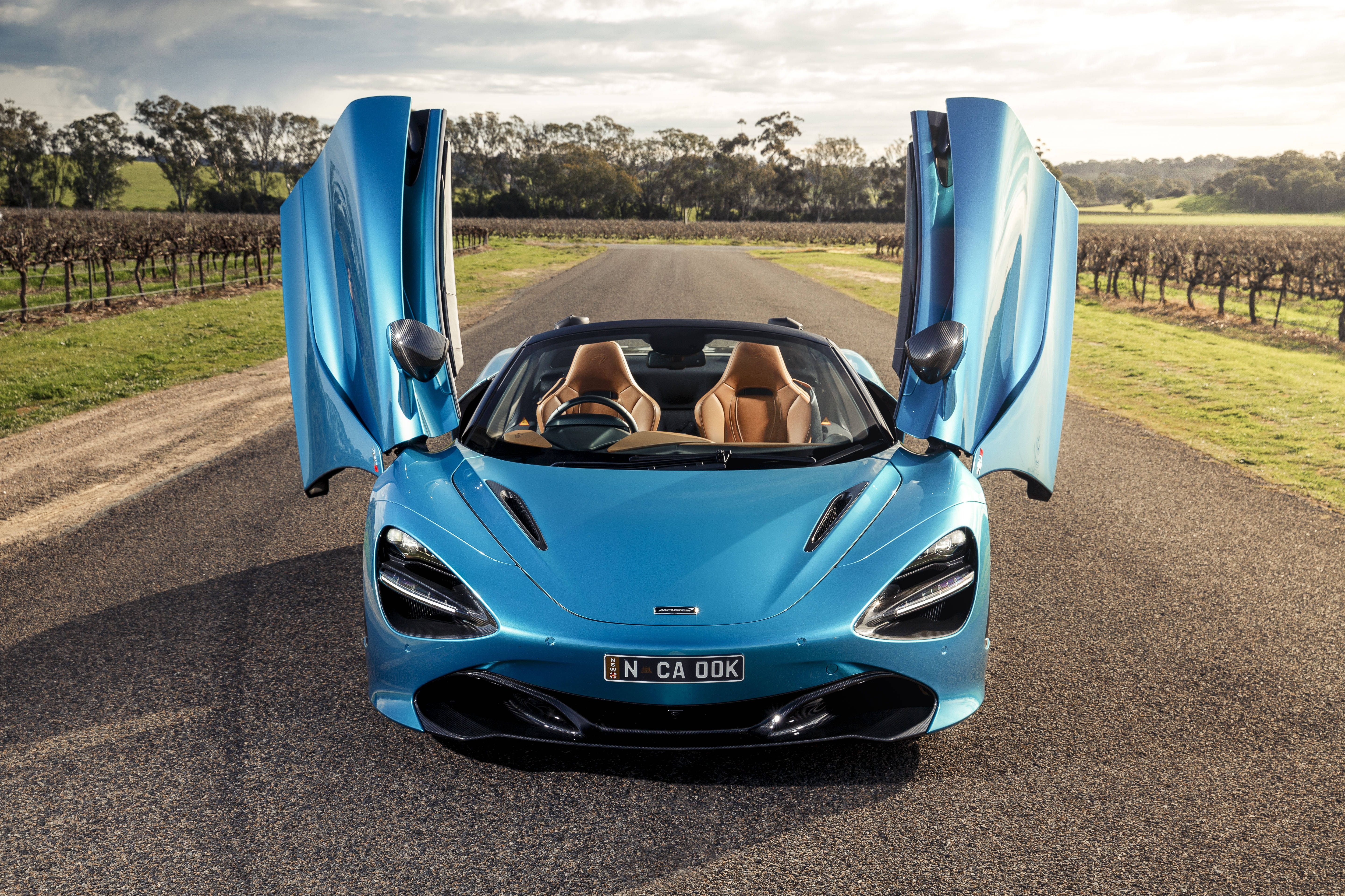 Mclaren 720s 桌布 壁紙 — 一輛鮮藍色McLaren 720S Spider打開鷗翼門，停在兩旁種滿葡萄園的鄉村道路上，天空柔和。