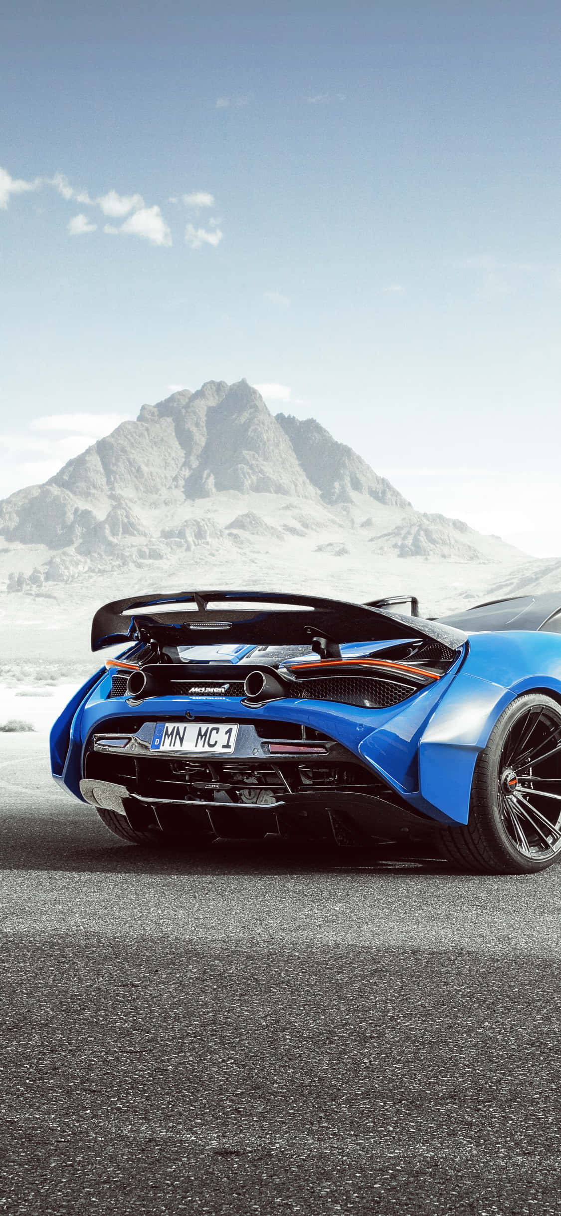 iPhone Xs Mclaren 720s 桌布 壁紙 — 一輛鮮藍色McLaren跑車，帶有大型後擾流板，停在瀝青路面上，背景是白雪覆蓋的山脈，天空晴朗。