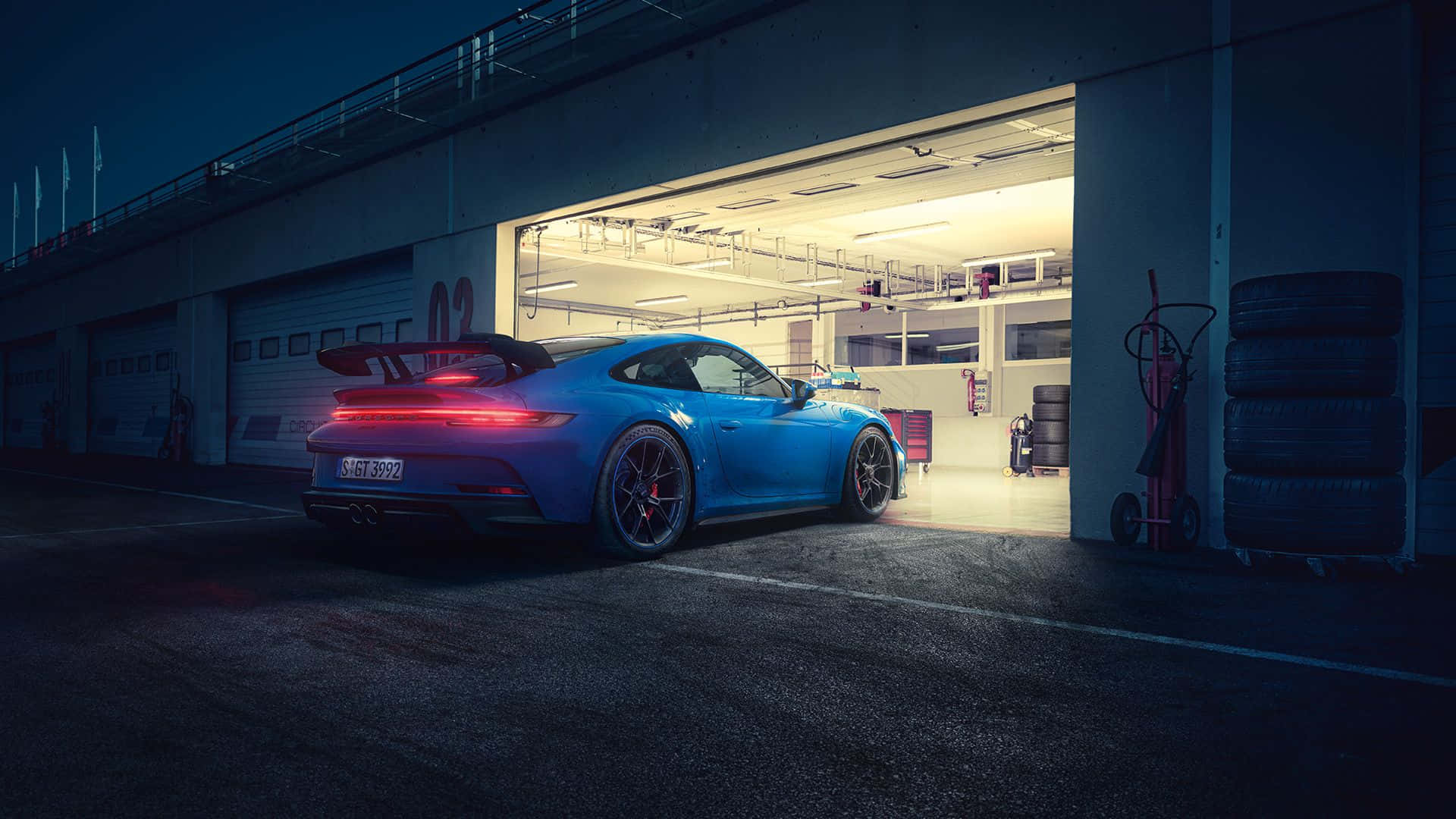 保時捷 桌布 壁紙 — 一輛鮮藍色保時捷 911 GT3 RS 在夜燈下閃閃發光，駛出一個裝滿賽車輪胎和設備的車庫。