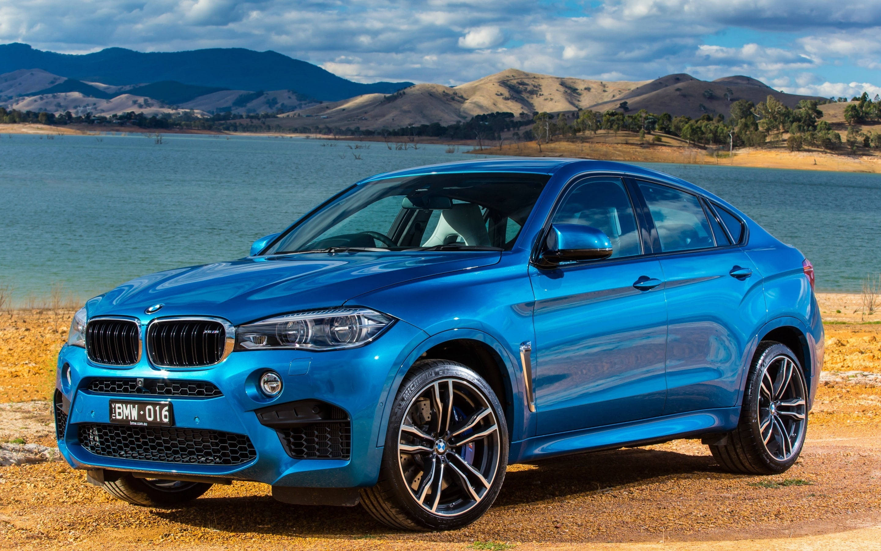 m 桌布 壁紙 — 一輛鮮藍色的BMW X6 M停在平靜湖泊旁的碎石上，背景有起伏的山丘和多雲的天空。