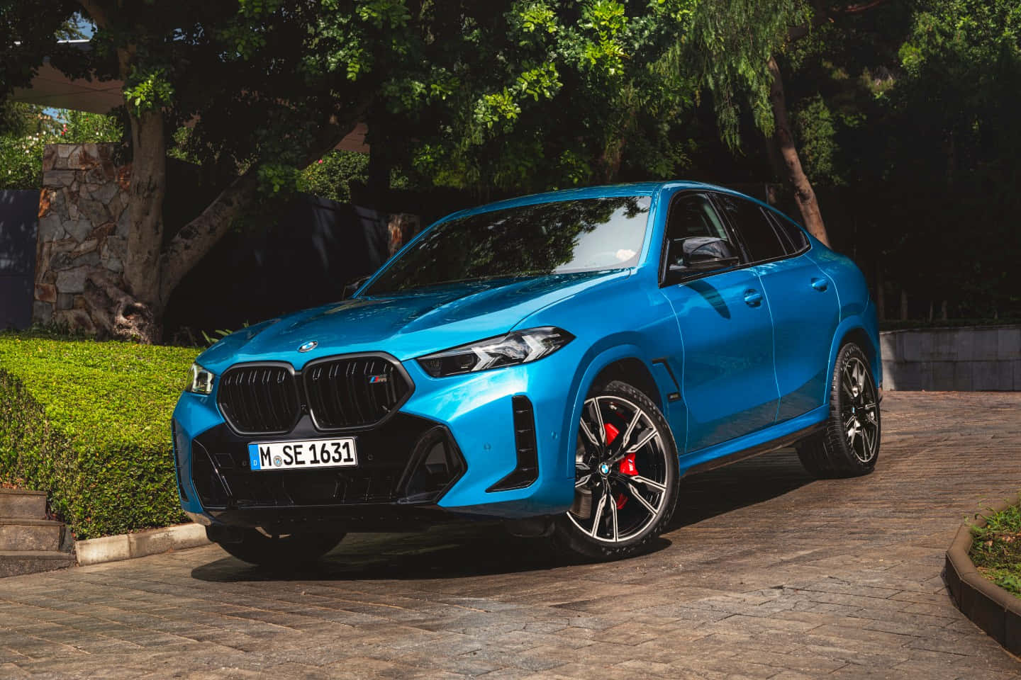 Bmw X5 桌布 壁紙 — 一輛鮮藍色的BMW X6 M停放在石頭車道上，周圍環繞著茂密的綠樹和修剪整齊的樹籬。