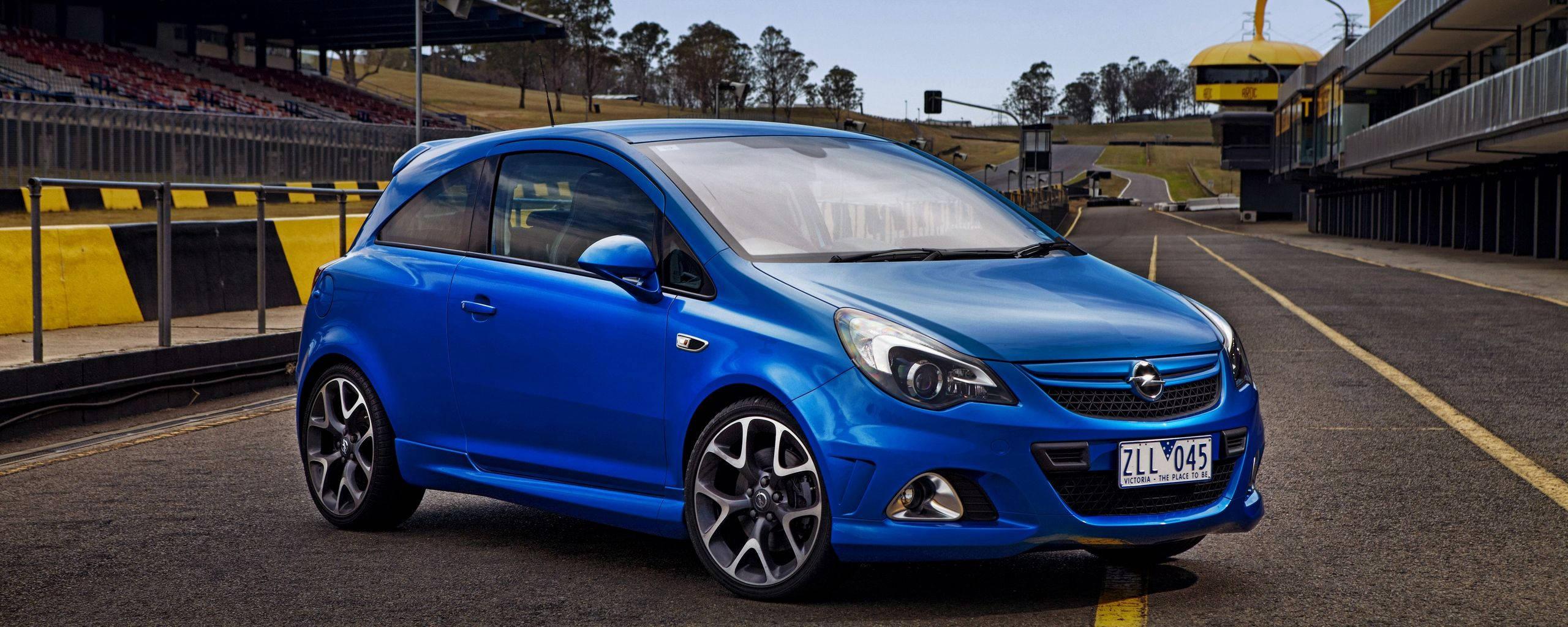 Opel 桌布 壁紙 — 一輛鮮藍色的歐寶雅特 GTC 跑車停泊在賽車場上，背景有看台和黃色護欄。