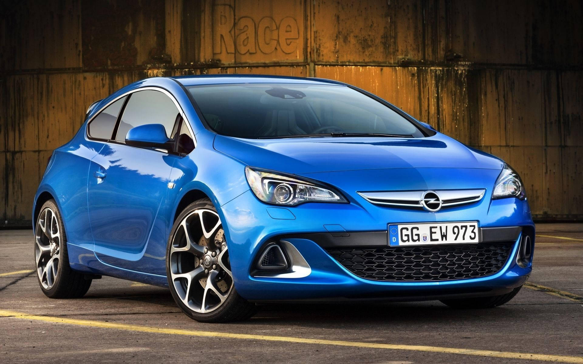 Opel 桌布 壁紙 — 一輛鮮藍色的歐寶雅特轎跑車在明亮燈光下閃閃發光，背景是刻有'Race'字樣的風化木牆。