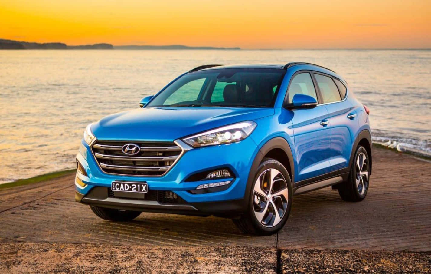 Hyundai 桌布 壁紙 — 一輛鮮藍色的現代 Tucson SUV 停泊在木製碼頭上，背景是日落時分的金色天空和寧靜的水面。