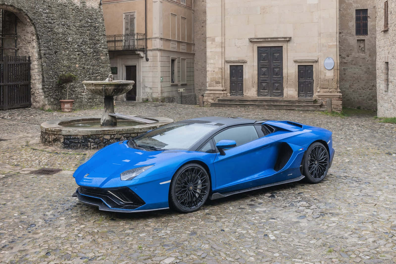 Lamborghini On Fire 桌布 壁紙 — 一輛鮮藍色的藍寶堅尼 Aventador 停泊在鵝卵石庭院，旁邊有石噴泉和歷史建築。