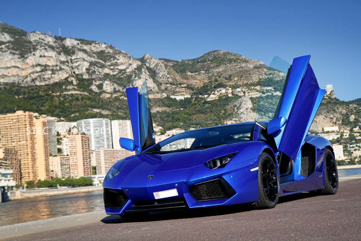 跑車 桌布 壁紙 — 一輛鮮藍色的藍寶堅尼Aventador，剪刀門打開，停泊在沿海公路上，背景是山脈和城市建築，天空清澈湛藍。
