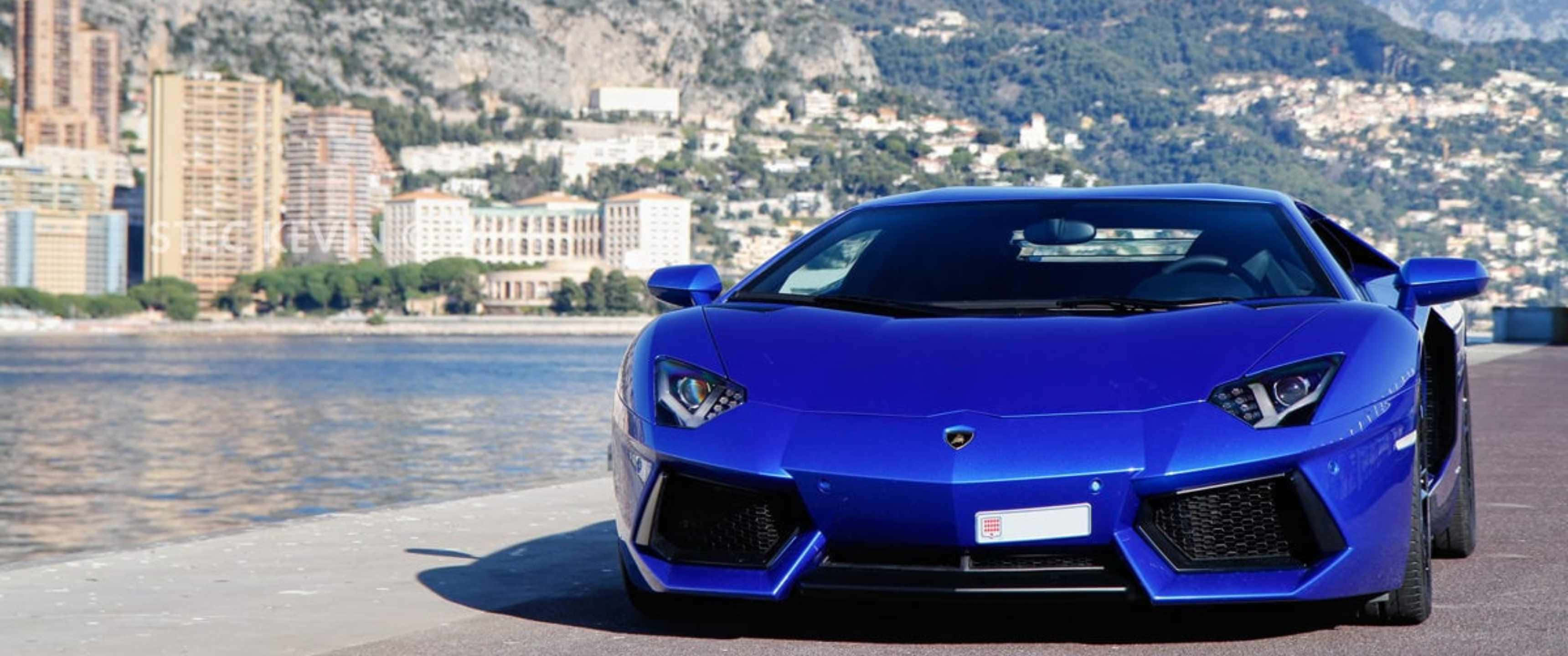 3440x1440p Lamborghini 桌布 壁紙 — 一輛鮮藍色藍寶堅尼Aventador停在海濱長廊上，背景是摩納哥的天際線和山脈。