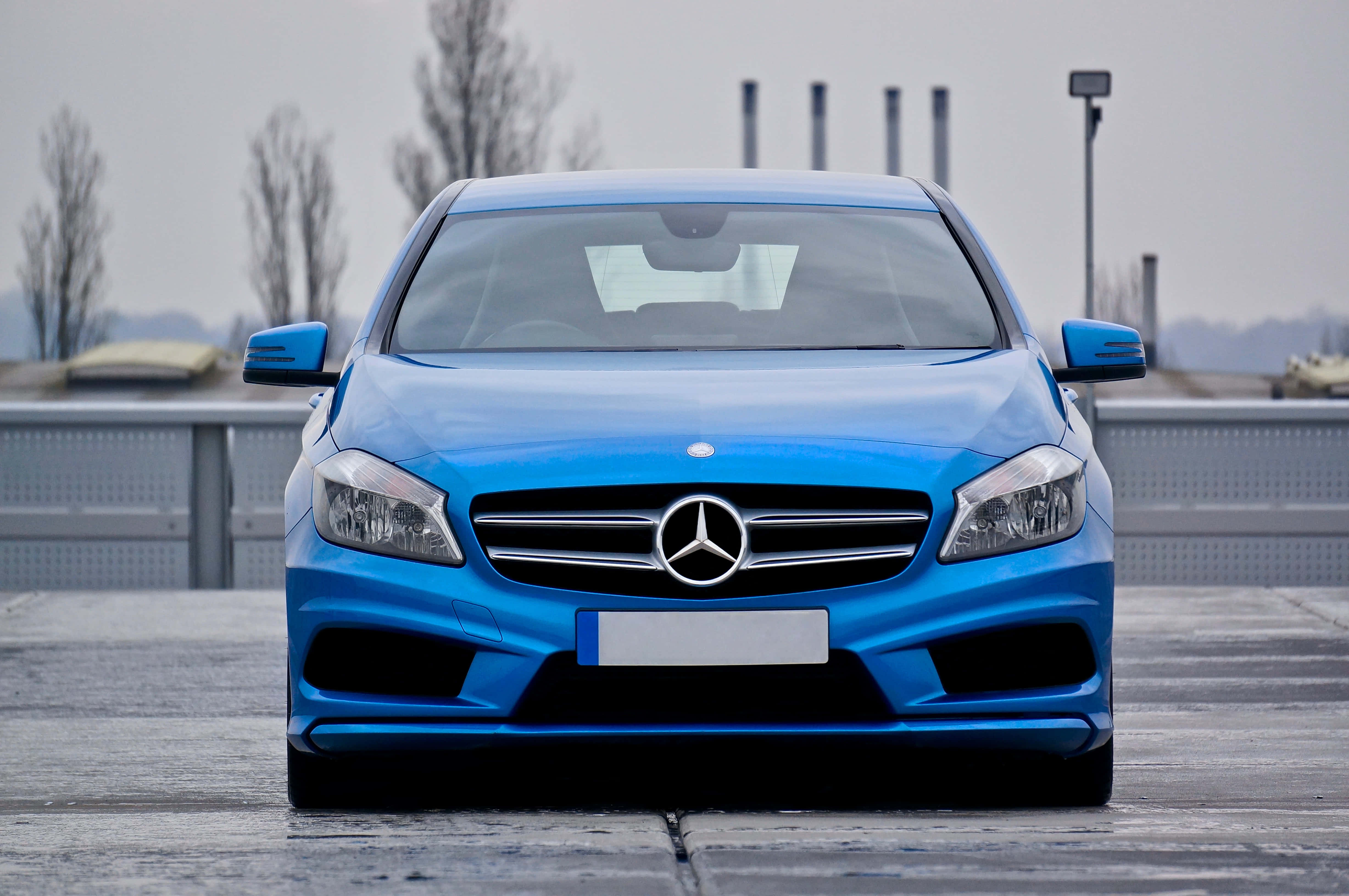 Mercedes Car 4K 桌布 壁紙 — 一輛鮮藍色賓士轎車面向前方，停在灰色混凝土表面上，背景陰天，有光禿樹木和工業建築。
