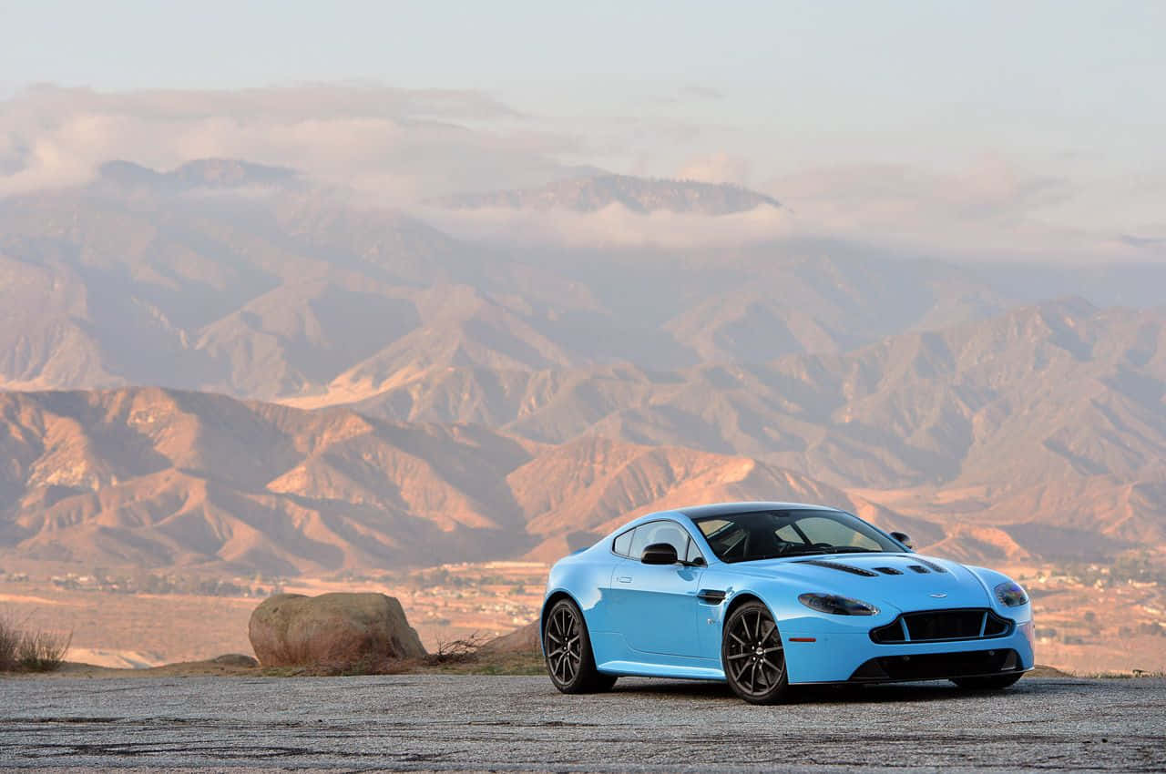 Aston Martin V12 Vantage 桌布 壁紙 — 一輛鮮藍色跑車停泊在沙漠觀景點，層層山脈在柔和日光下逐漸消失在霧靄中。