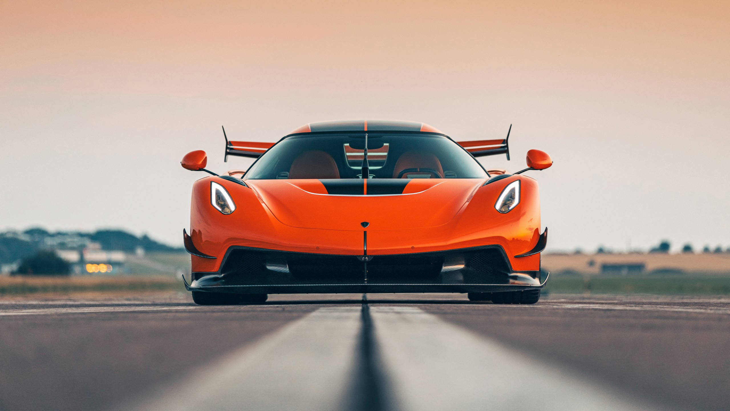 跑車 桌布 壁紙 — 一輛鮮豔的橙色 Koenigsegg 超跑在日落時分面向跑道，天空呈現柔和漸變色。