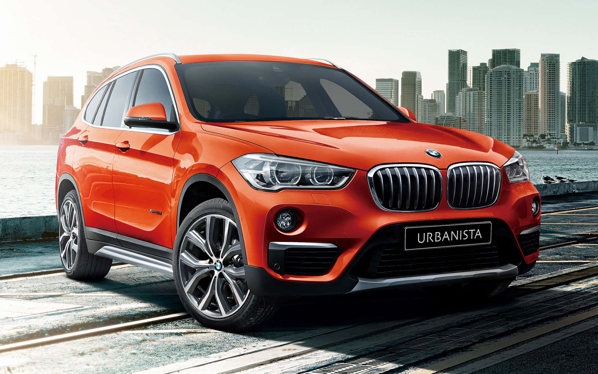 Bmw X1 桌布 壁紙 — 一輛鮮豔的橙色BMW SUV停在海濱長廊上，背景是現代化的城市天際線。