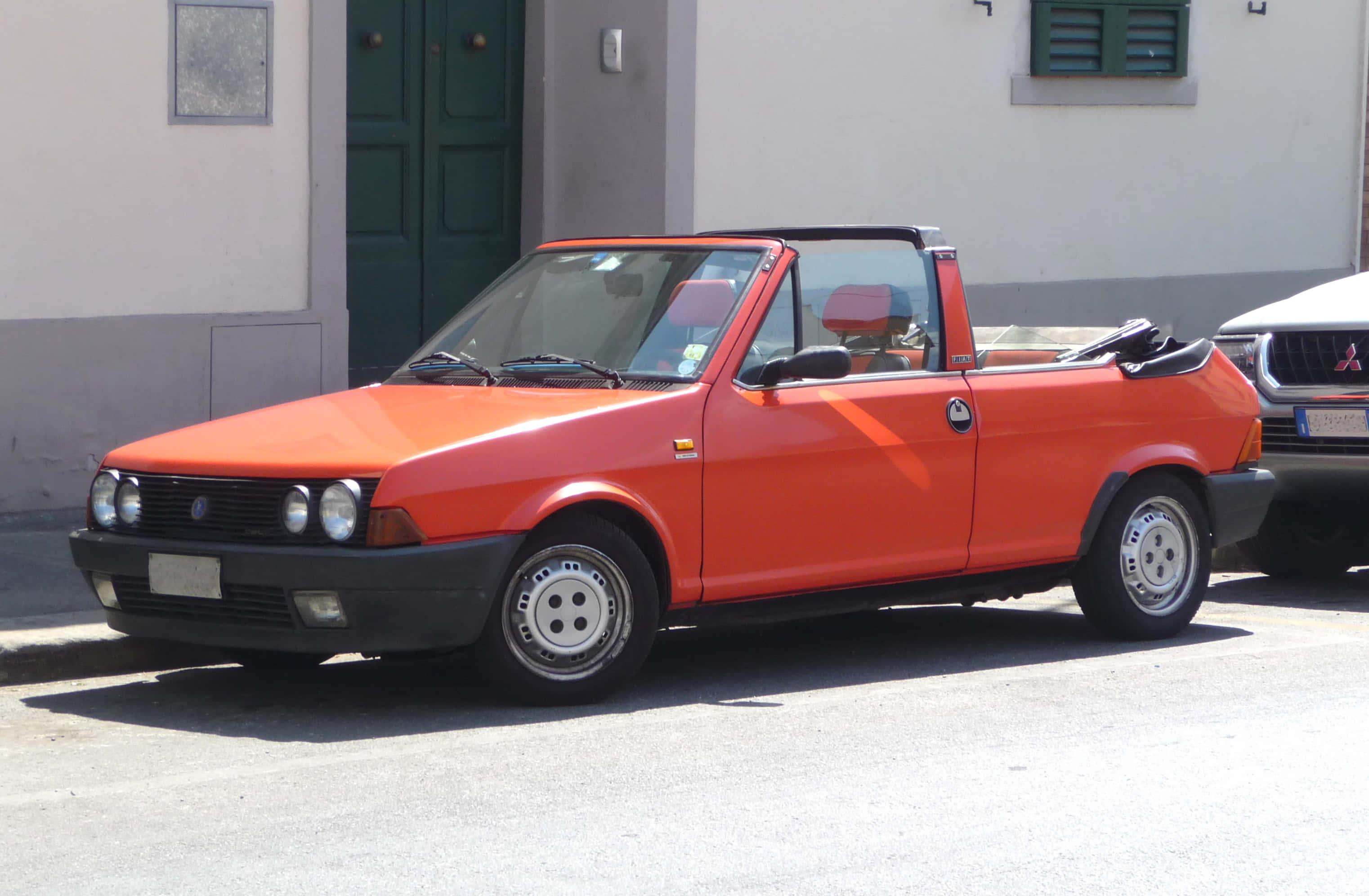 Fiat Ritmo 桌布 壁紙 — 一輛鮮豔的橙色Fiat 126 Spider敞篷車停放在陽光明媚的街道上，旁邊是一棟有綠色百葉窗的建築物。