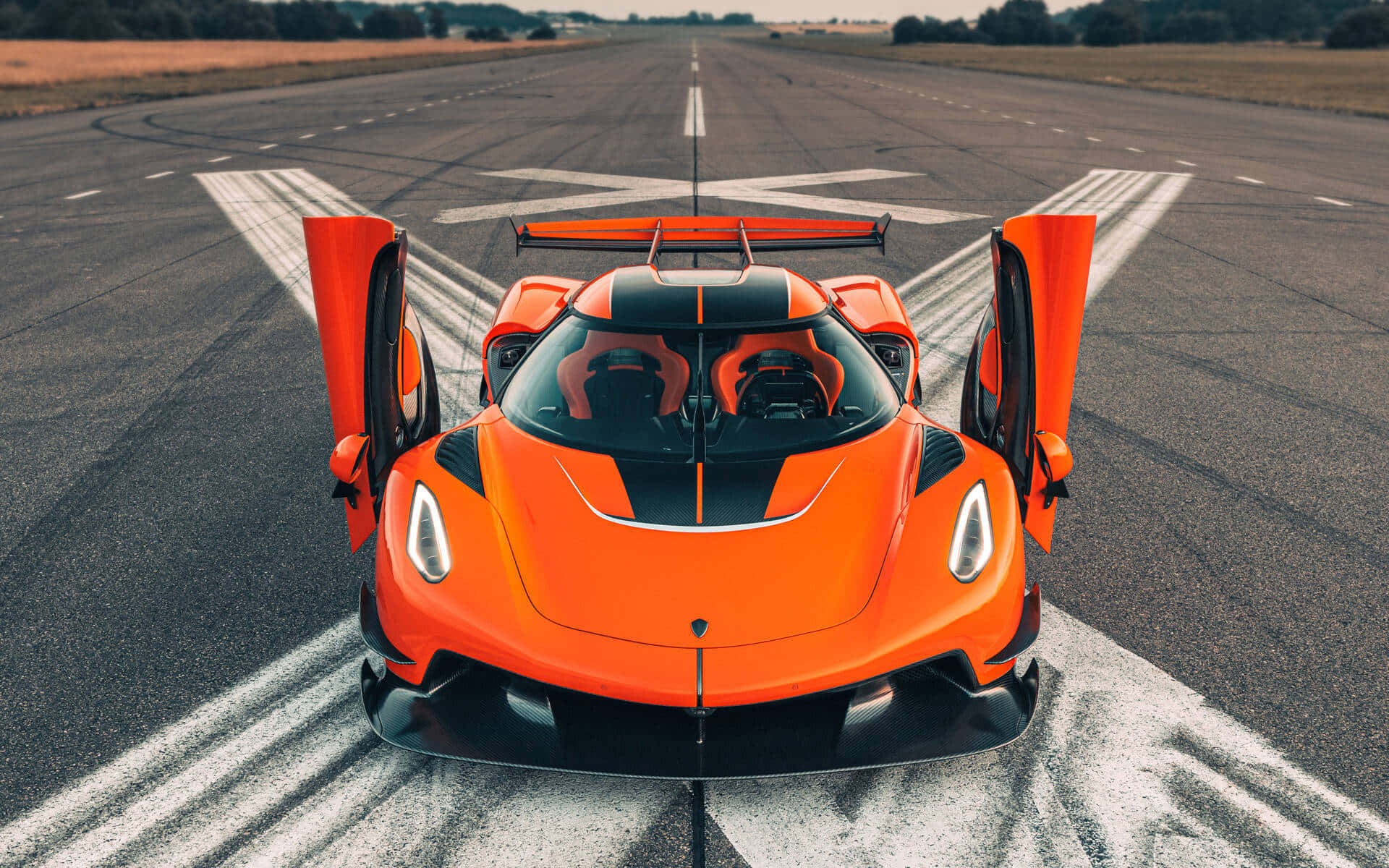 Koenigsegg 桌布 壁紙 — 一輛鮮豔的橙色Koenigsegg超跑打開車門，停在一條有白色標線的寬闊跑道中央，在晴朗的日光下。