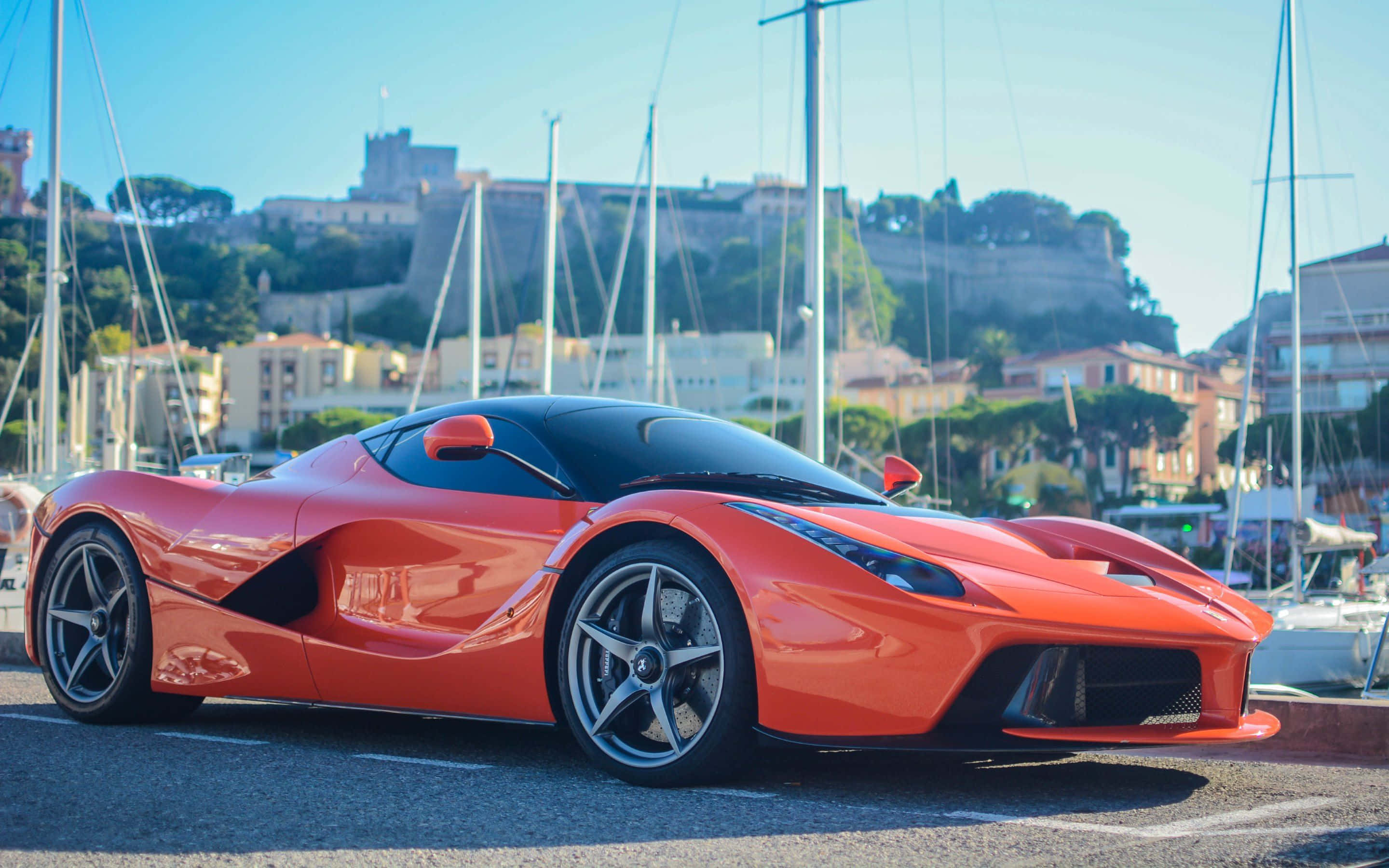Ferrari Laferrari 桌布 壁紙 — 一輛鮮豔的橙色LaFerrari超級跑車在明亮的陽光下閃閃發光，停泊在一個遊艇碼頭，背景有帆船和歷史建築。
