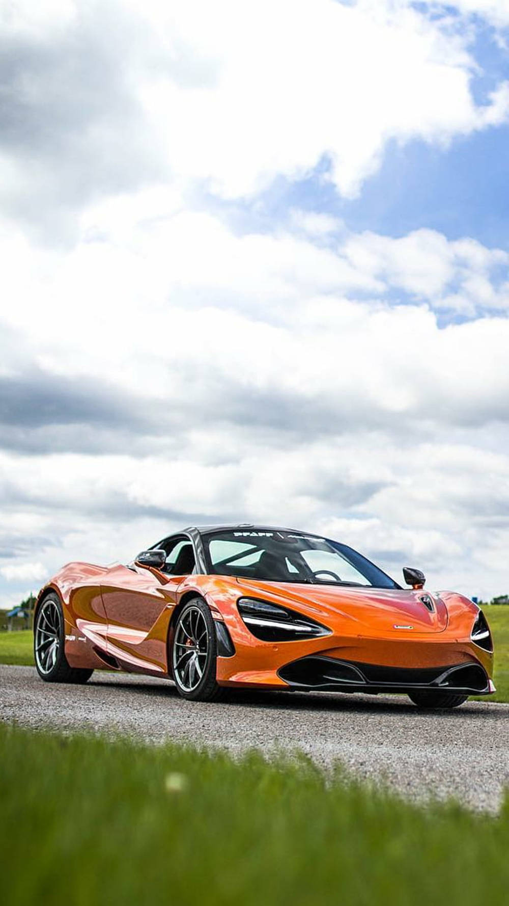 Grass 桌布 壁紙 — 一輛鮮豔的橙色McLaren 720S跑車停泊在碎石路上，背景是戲劇性的多雲天空，前景有綠草。