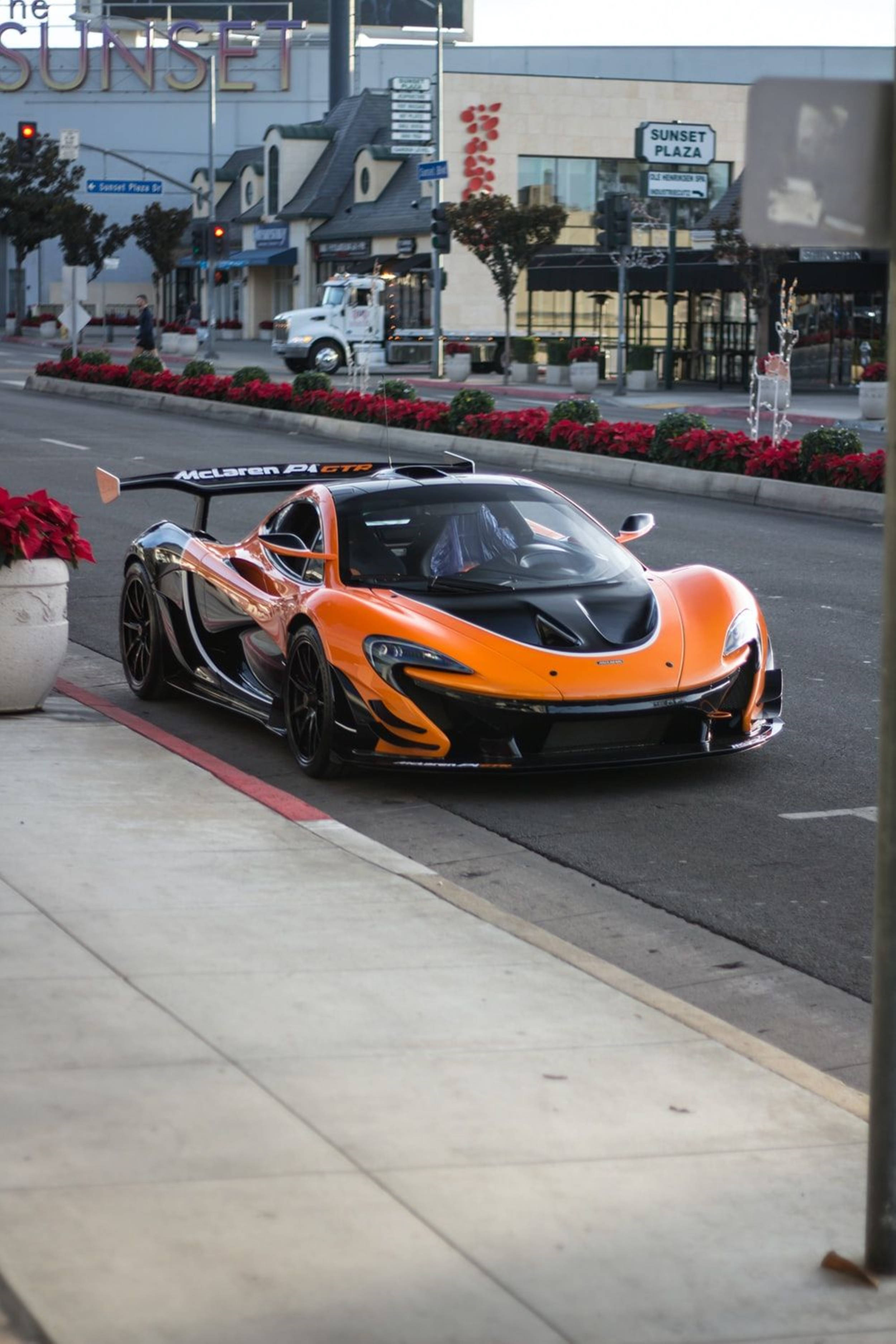 street photography 桌布 壁紙 — 一輛鮮豔的橙色McLaren P1 GTR跑車停放在城市的街道上，旁邊有紅色聖誕紅和Sunset Plaza標誌。
