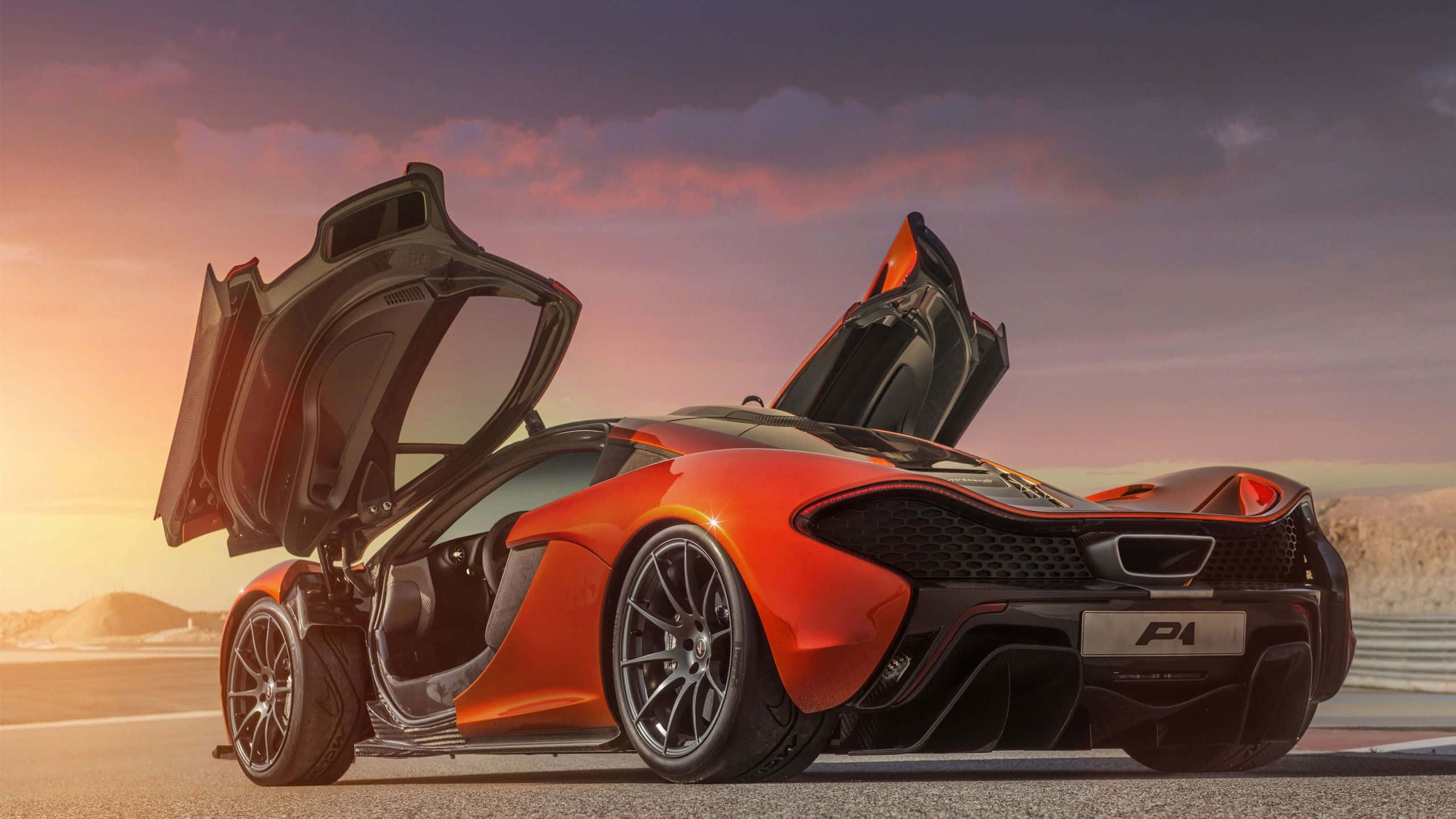 跑車 桌布 壁紙 — 一輛鮮豔的橙色McLaren P1超級跑車，鷗翼車門打開，停在夕陽下的賽道上。