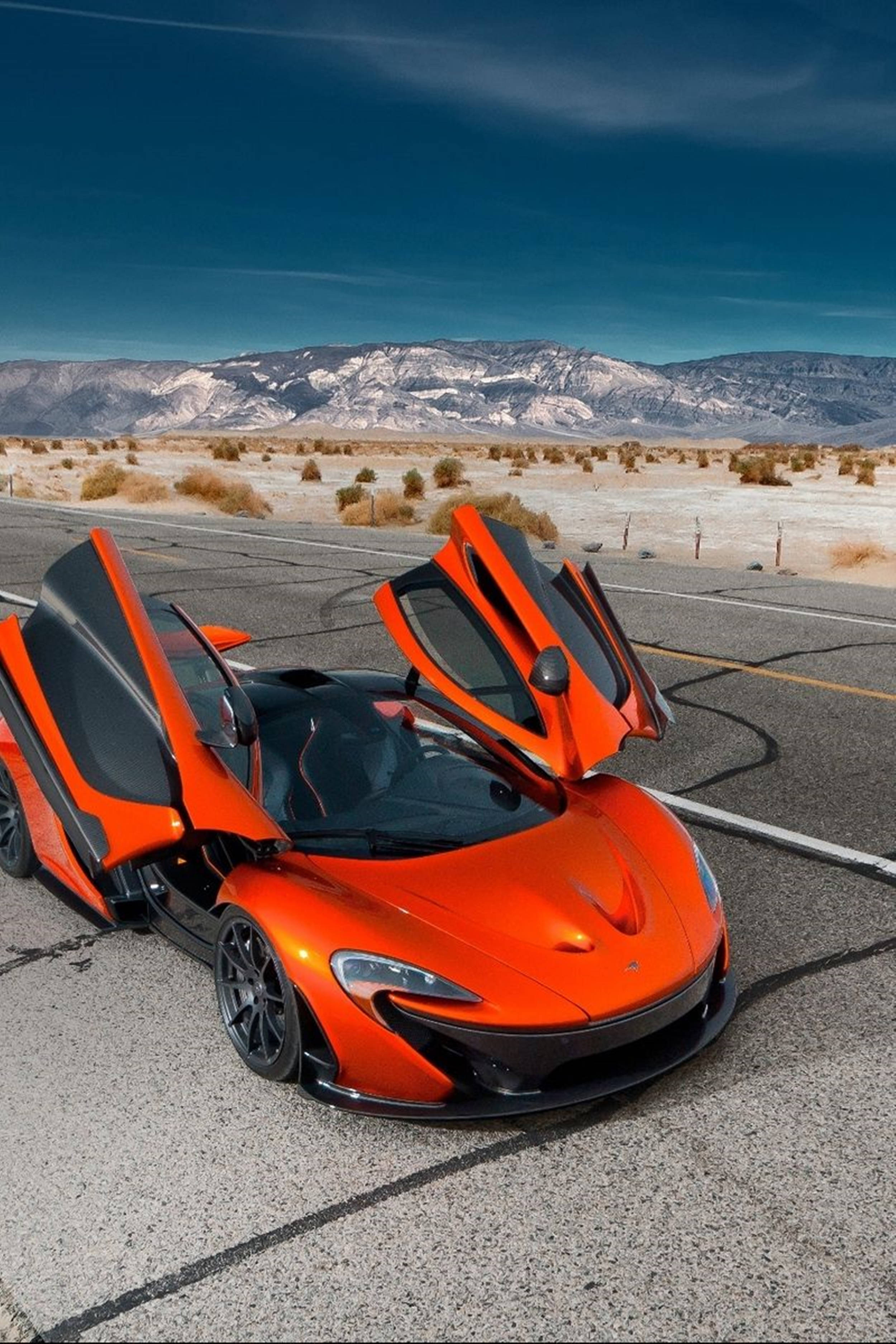 Luxury 桌布 壁紙 — 一輛鮮豔的橙色McLaren跑車，蝴蝶門打開，停在沙漠公路上，天空湛藍，遠處有積雪覆蓋的山脈。
