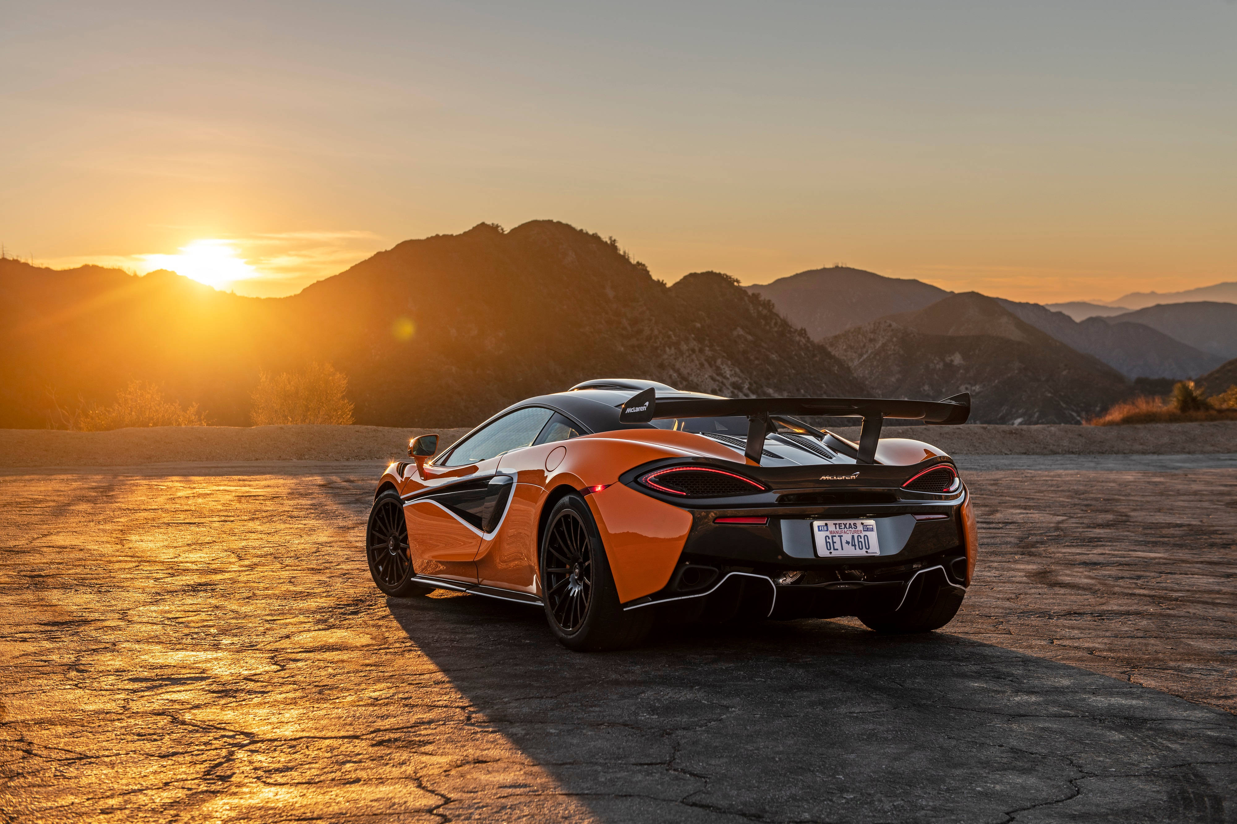 Auto Racing 桌布 壁紙 — 一輛鮮豔的橙色McLaren跑車停在裂開的路面上，背景有山脈和金黃色的日落。