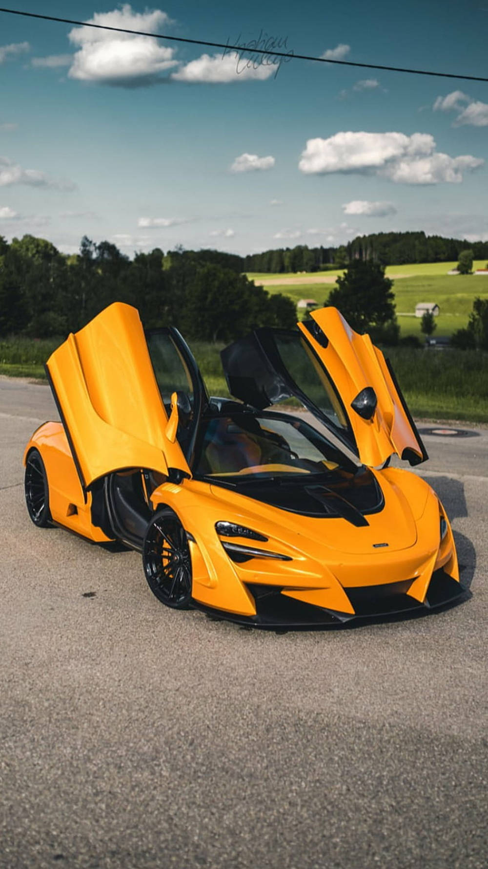 跑車 桌布 壁紙 — 一輛鮮豔的橙色McLaren跑車，鷗翼車門打開，停在鄉村道路上，上方是晴朗的藍天和蓬鬆的白雲。