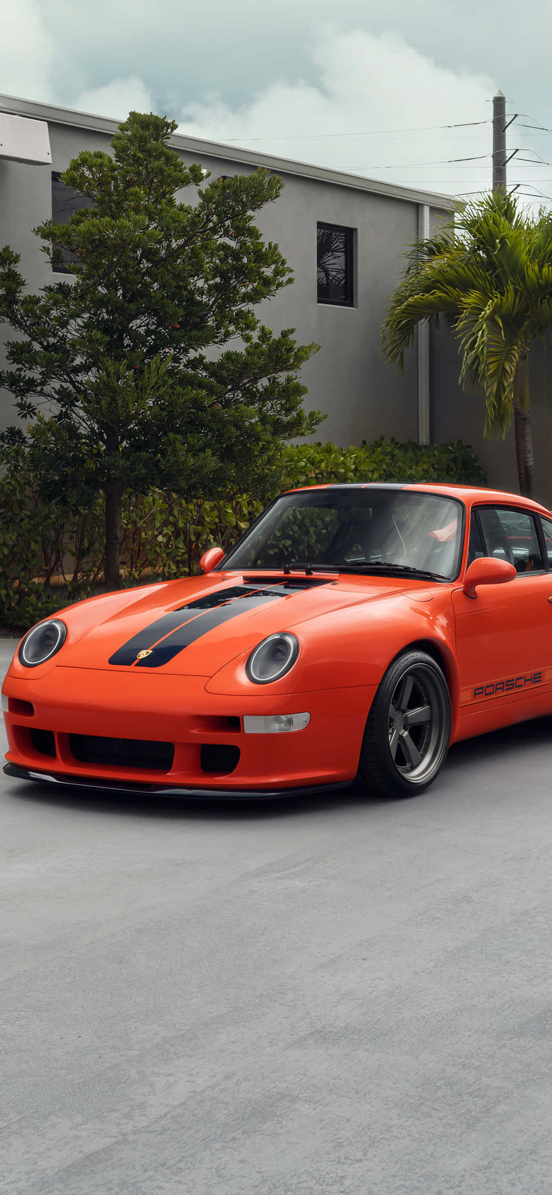 Porsche iPhone 桌布 壁紙 — 一輛鮮豔的橙色保時捷跑車，帶有黑色賽車條紋，停在混凝土車道上，旁邊是茂密的綠樹和一座現代化的灰色建築，天空多雲。