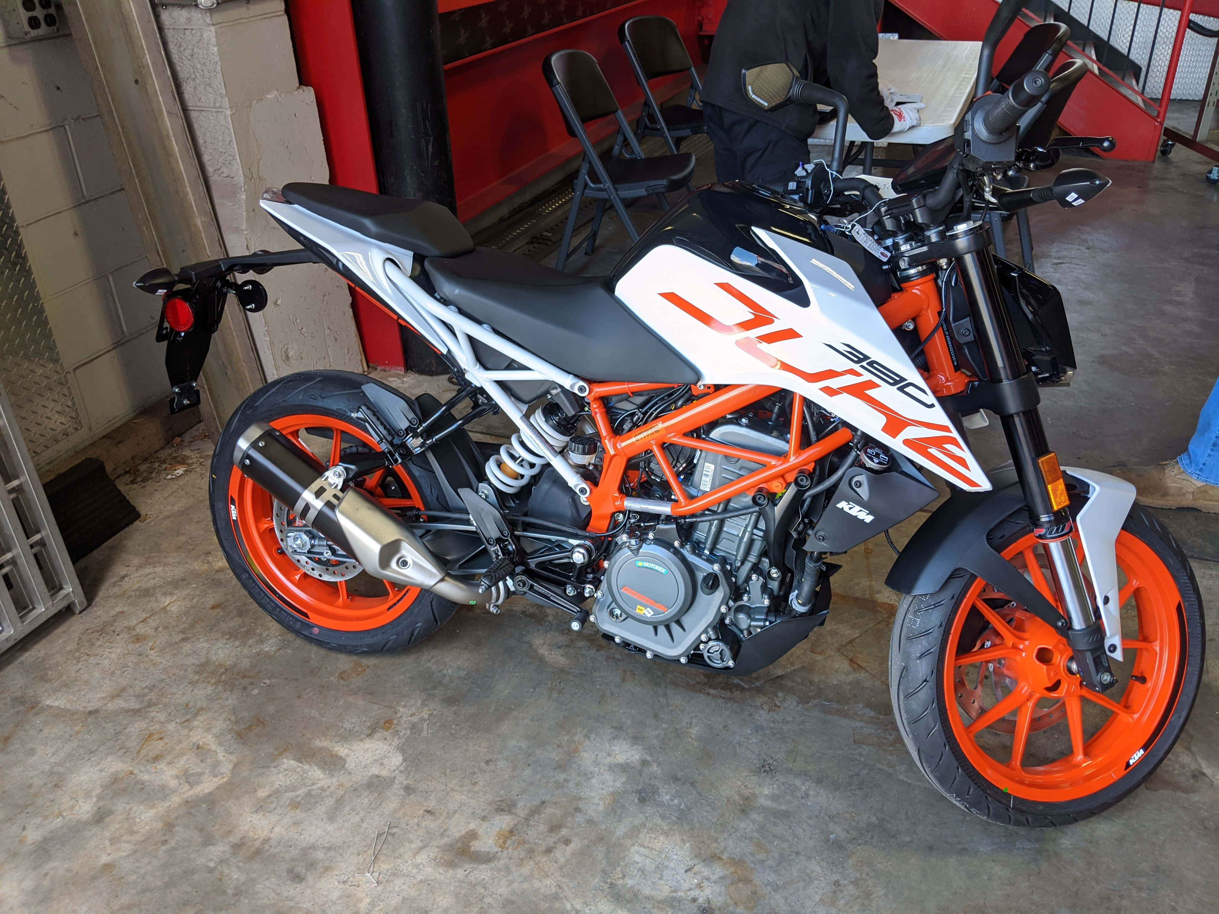 Garage 桌布 壁紙 — 一輛鮮豔的橙色和白色KTM Duke 390摩托車停放在室內混凝土地板上，背景中有一個人。