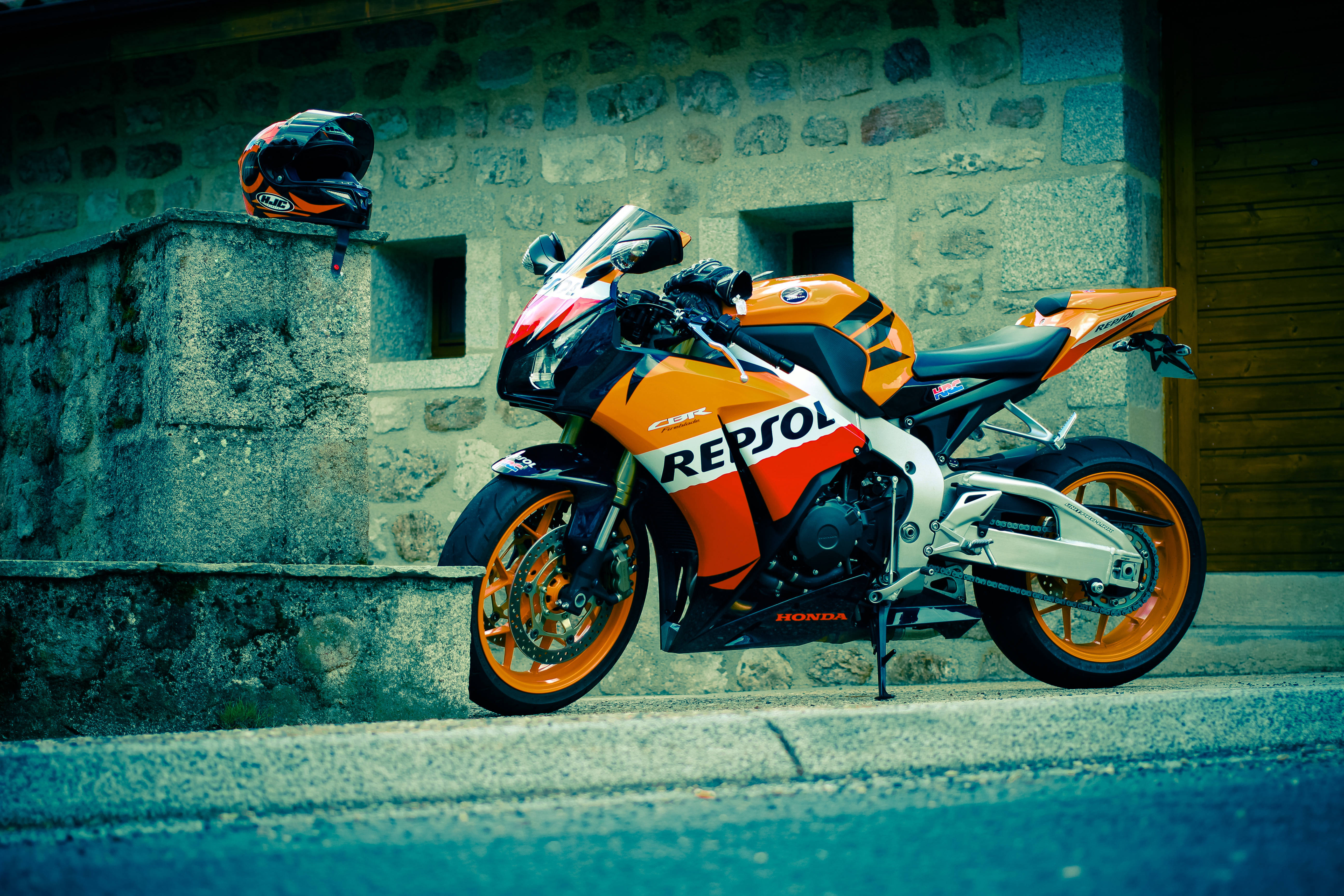 本田 桌布 壁紙 — 一輛鮮豔的橙色和白色本田 Repsol 賽車機車停放在石牆旁邊，上面放著一個相配的安全帽。