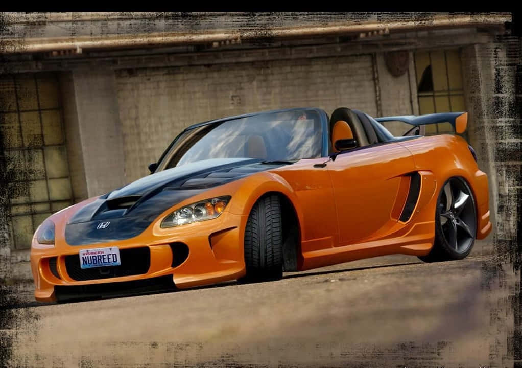Honda S2000 桌布 壁紙 — 一輛鮮豔的橙色和黑色本田S2000跑車，帶有客製化尾翼，停在風化的磚砌建築前。