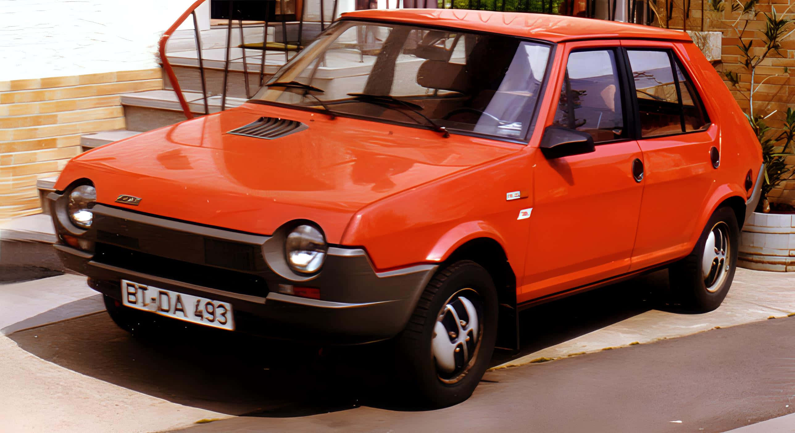 Fiat Ritmo 桌布 壁紙 — 一輛鮮豔的橙色復古菲亞特熊貓停放在陽光普照的人行道上，旁邊是磚砌建築和樓梯。