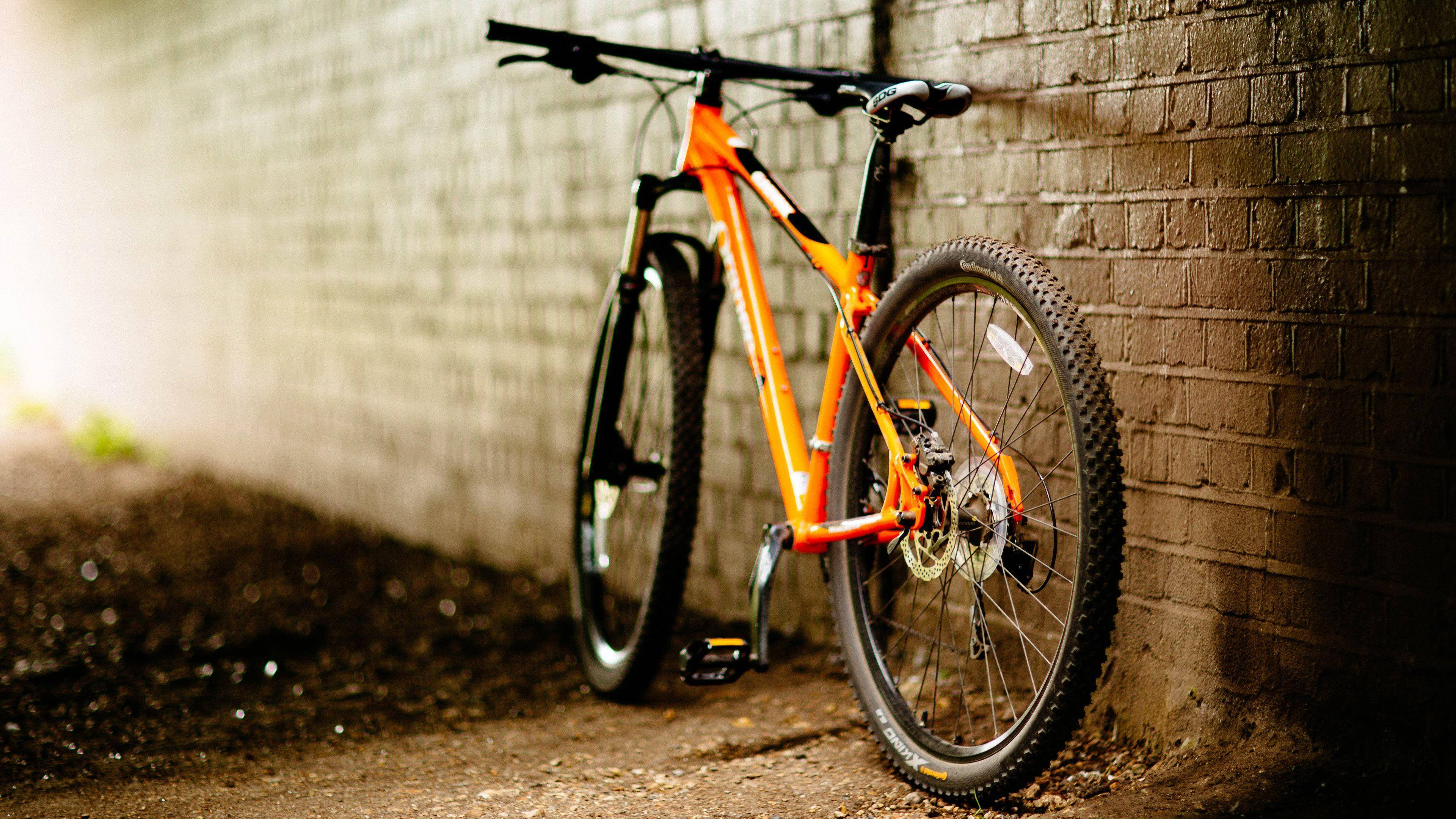 4K bike 桌布 壁紙 — 一輛鮮豔的橙色登山車靠在一面風化的磚牆上，柔和的光線從左側照射進來。