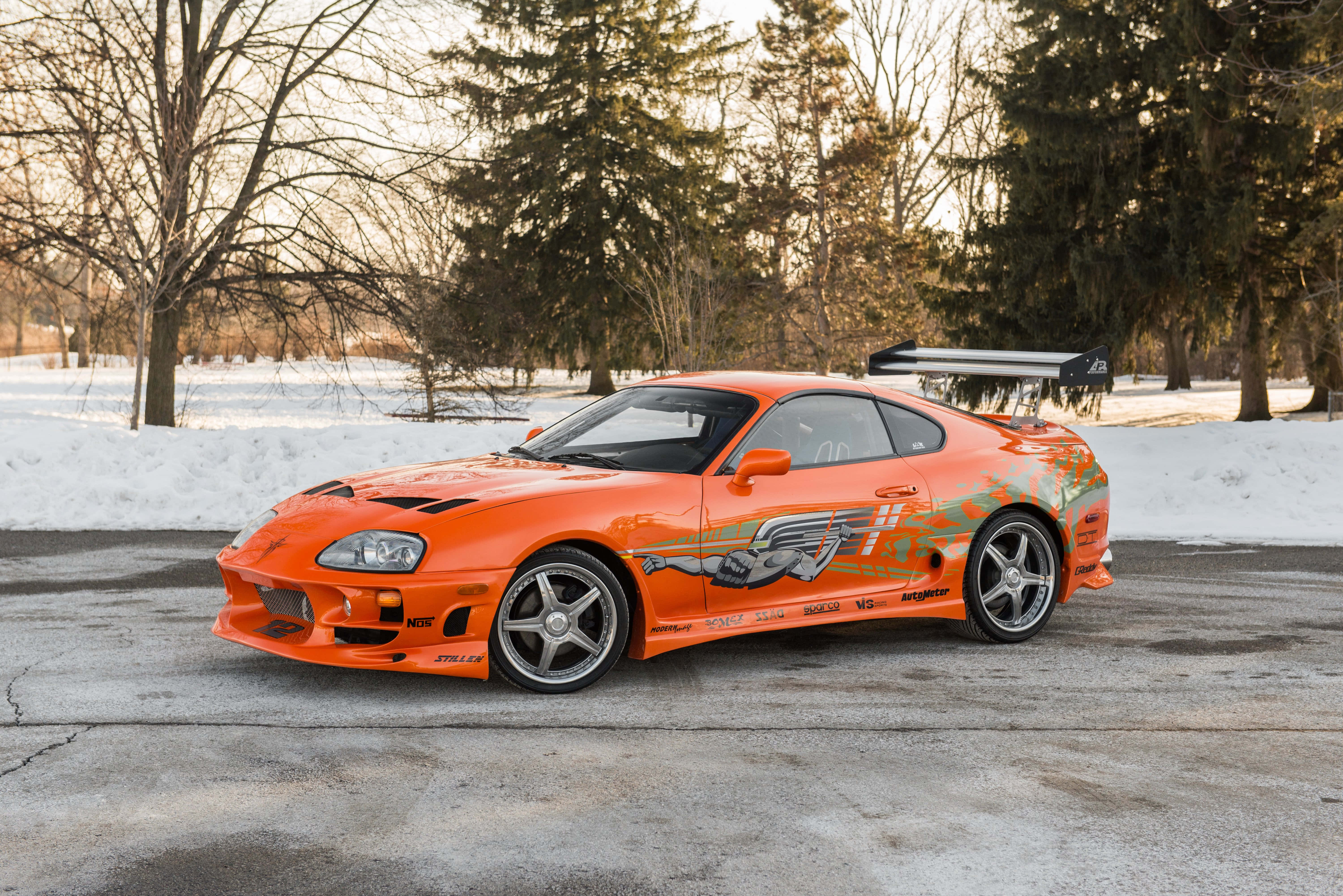 Fast And Furious 1 桌布 壁紙 — 一輛鮮豔的橙色豐田Supra帶有賽車貼紙，在黃金時段停泊在雪地車道上。